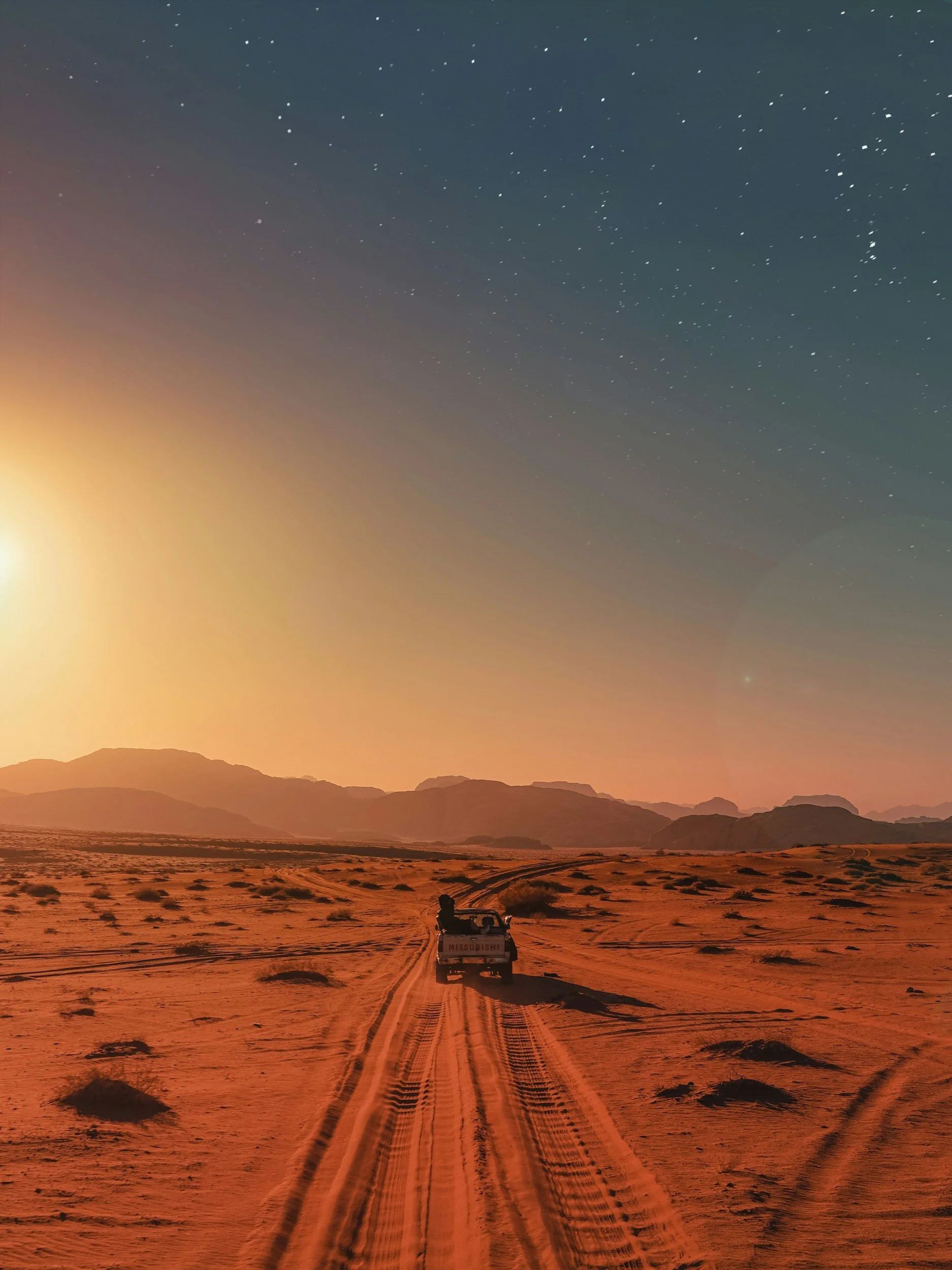 A vehicle driving on a sandy desert road at sunset with mountains in the background and a starry sky overhead.