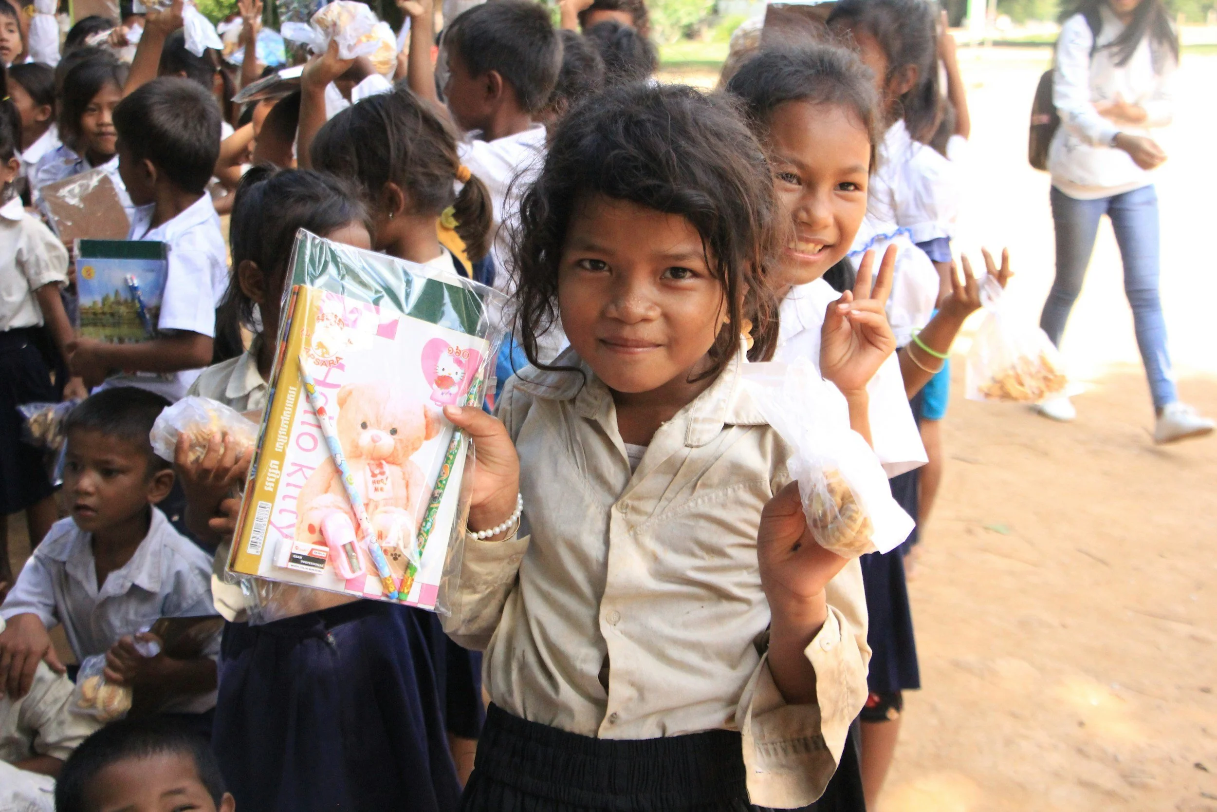 Children in school uniforms standing outdoors, holding school supplies and making peace signs, with a young girl in the foreground smiling.