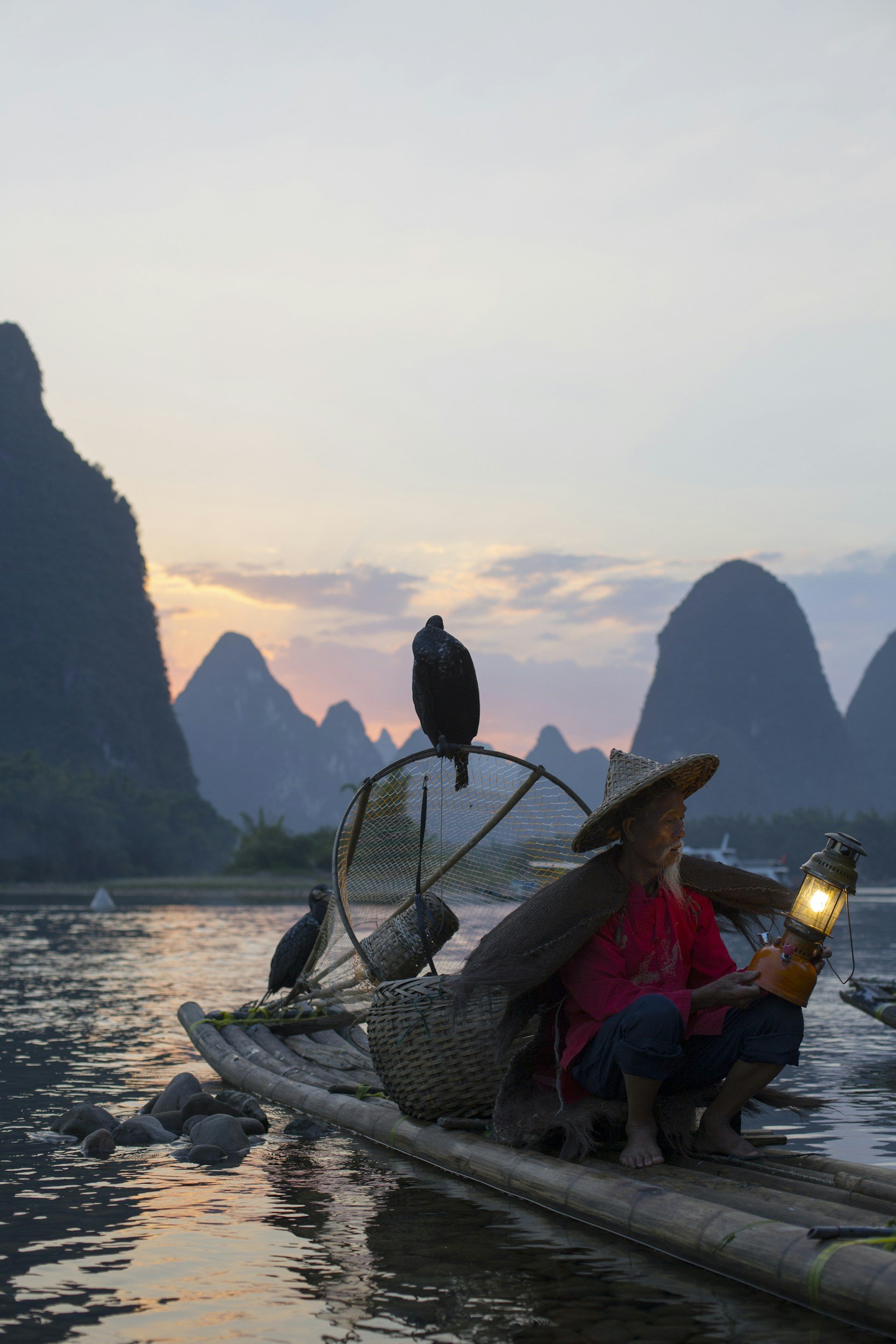 Elderly woman in traditional attire sitting on bamboo raft at riverside during sunset, holding a lantern, with karst mountains in the background.