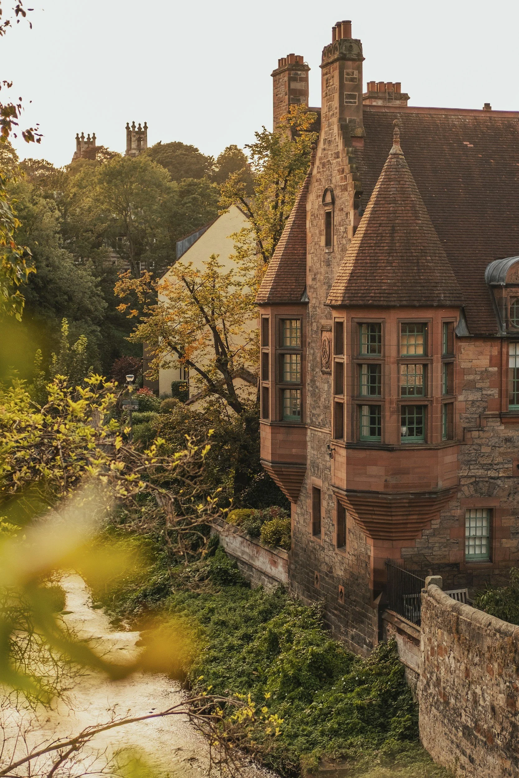 A historic stone house with a turret and multiple chimneys, situated beside a creek surrounded by greenery and trees, with additional historic chimneys visible on the distant horizon.