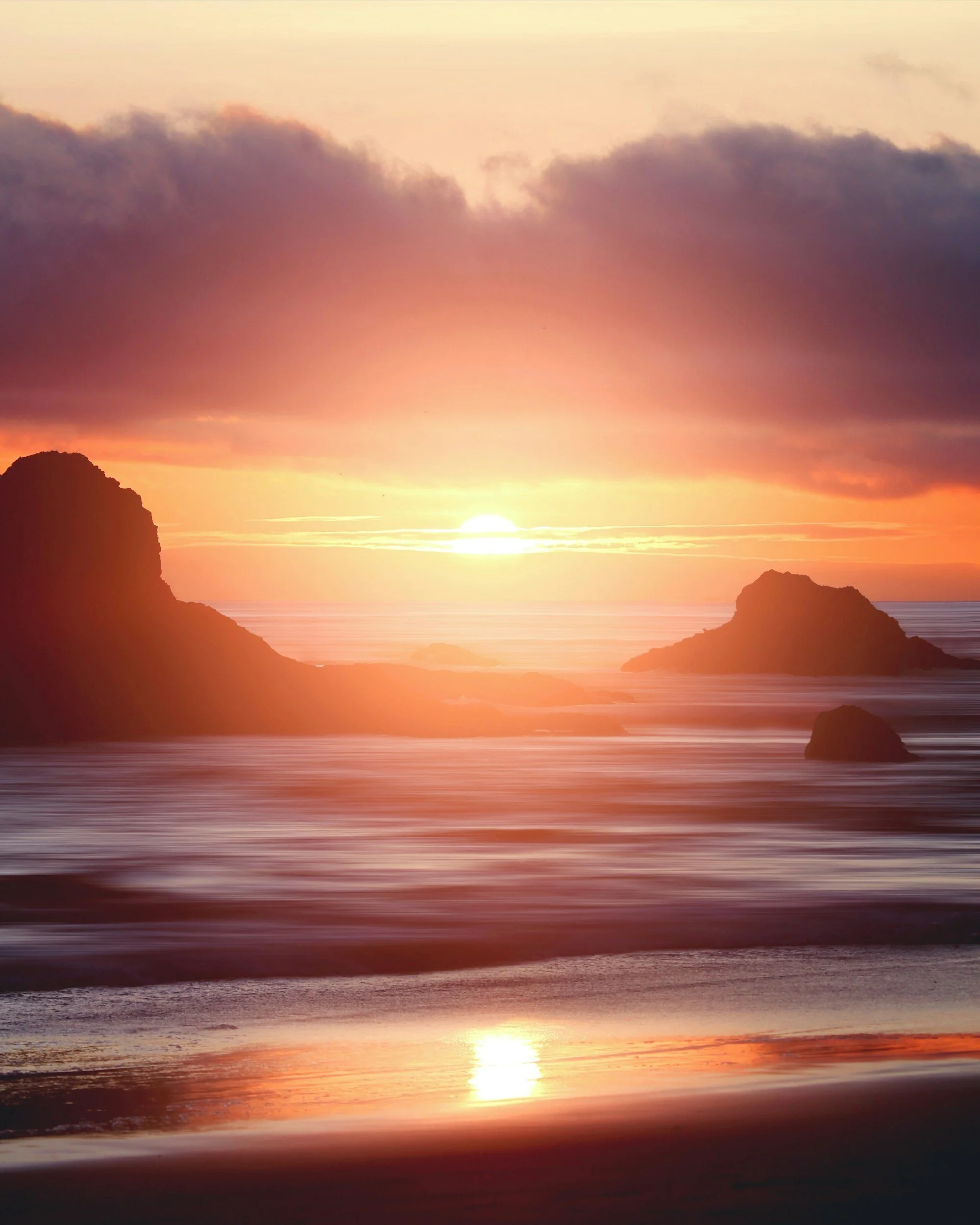 A scenic sunset over the ocean with the sun near the horizon, illuminating the sky with warm colors, and silhouetted rocky formations on both sides of the image.
