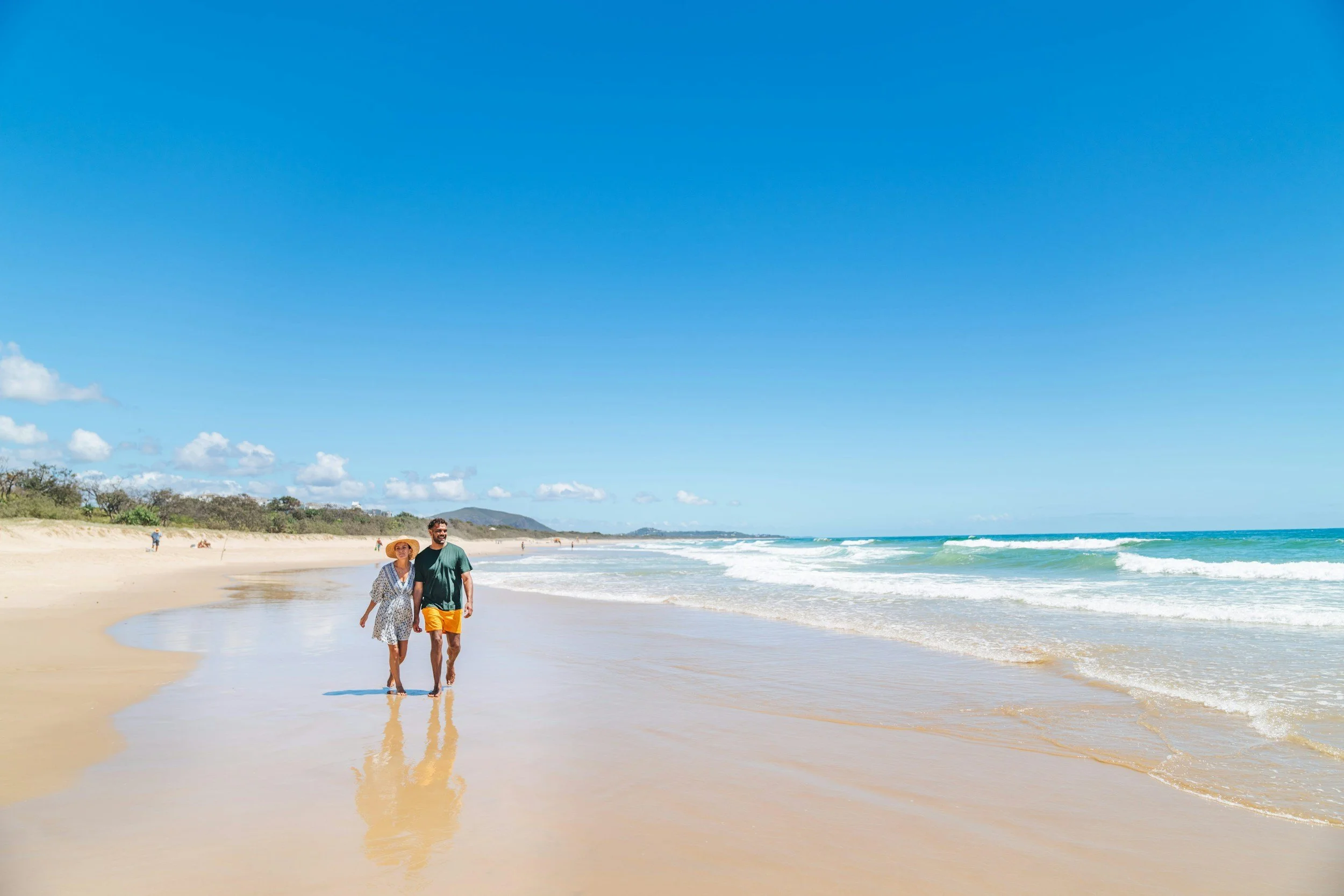 A couple walking along the shoreline of a sandy beach with blue sky and ocean waves.
