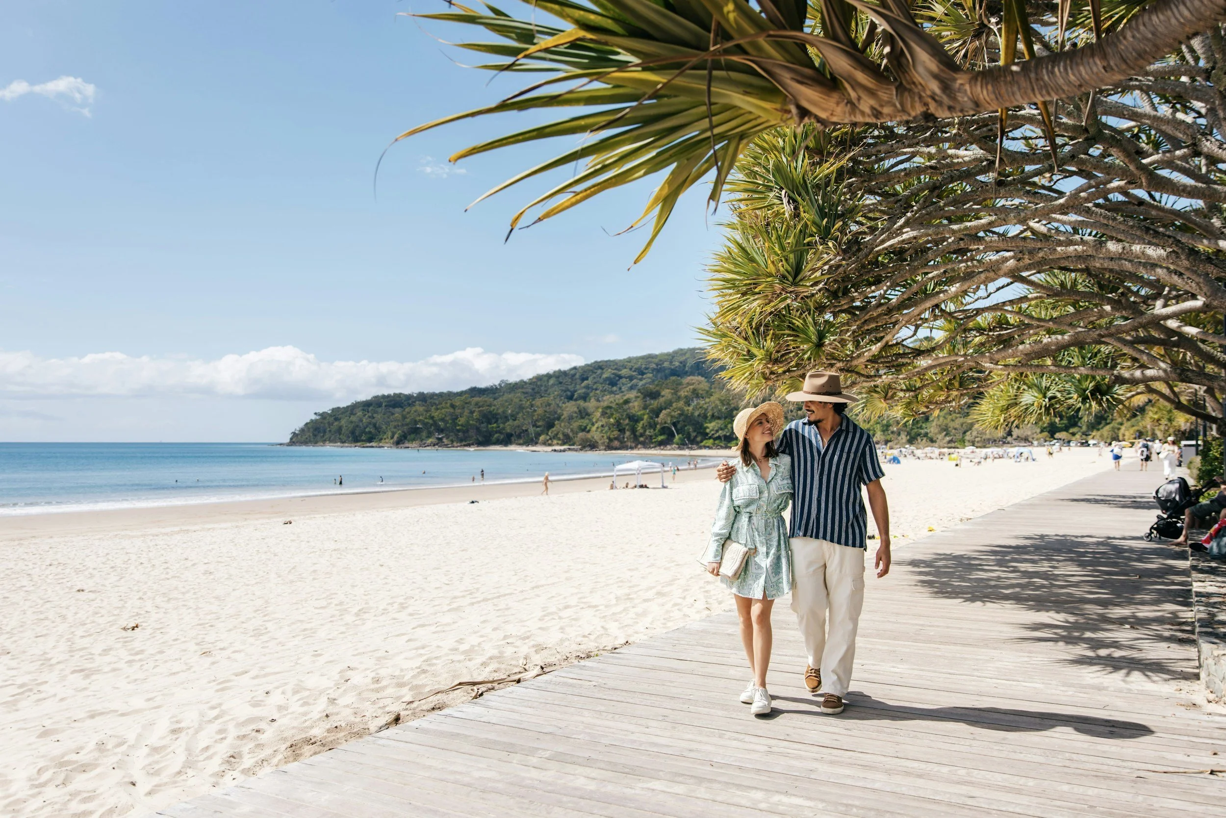 A couple walking along a beach boardwalk under large tropical trees, with the ocean and distant hills in the background. The woman is wearing a hat and a light dress, and the man is wearing a striped shirt and a wide-brim hat, smiling at each other.