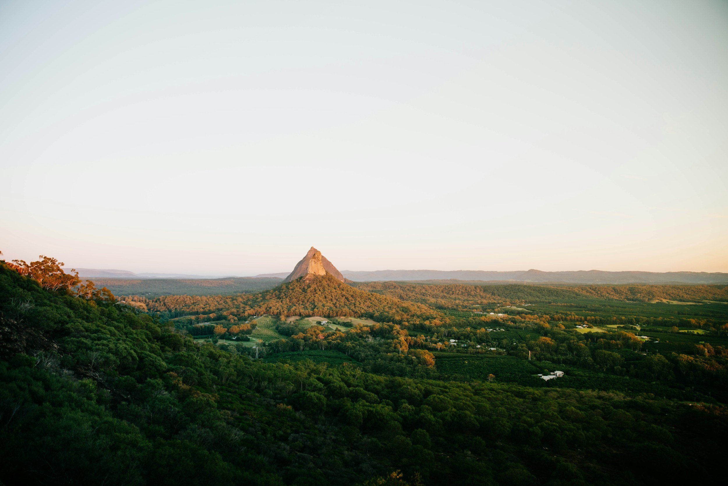 A landscape with a prominent tall, triangular mountain in the background, surrounded by green forested hills and open fields, under a clear sky.