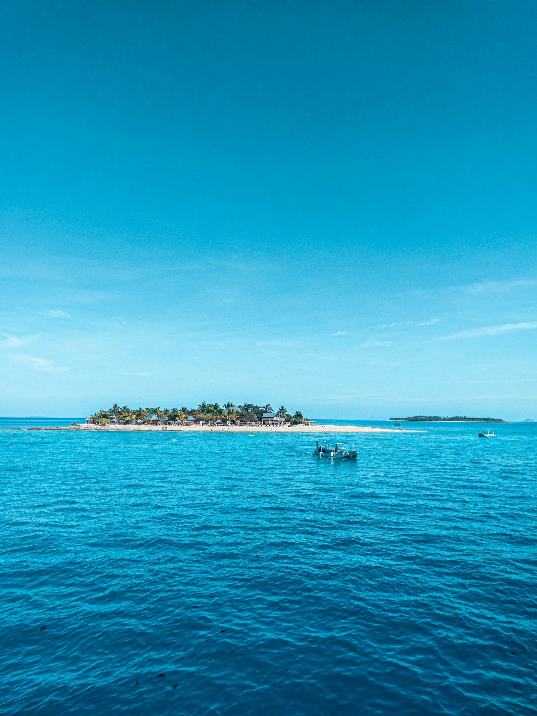 A scenic view of a small island with palm trees, buildings, and a sandy beach surrounded by turquoise water under a clear blue sky.