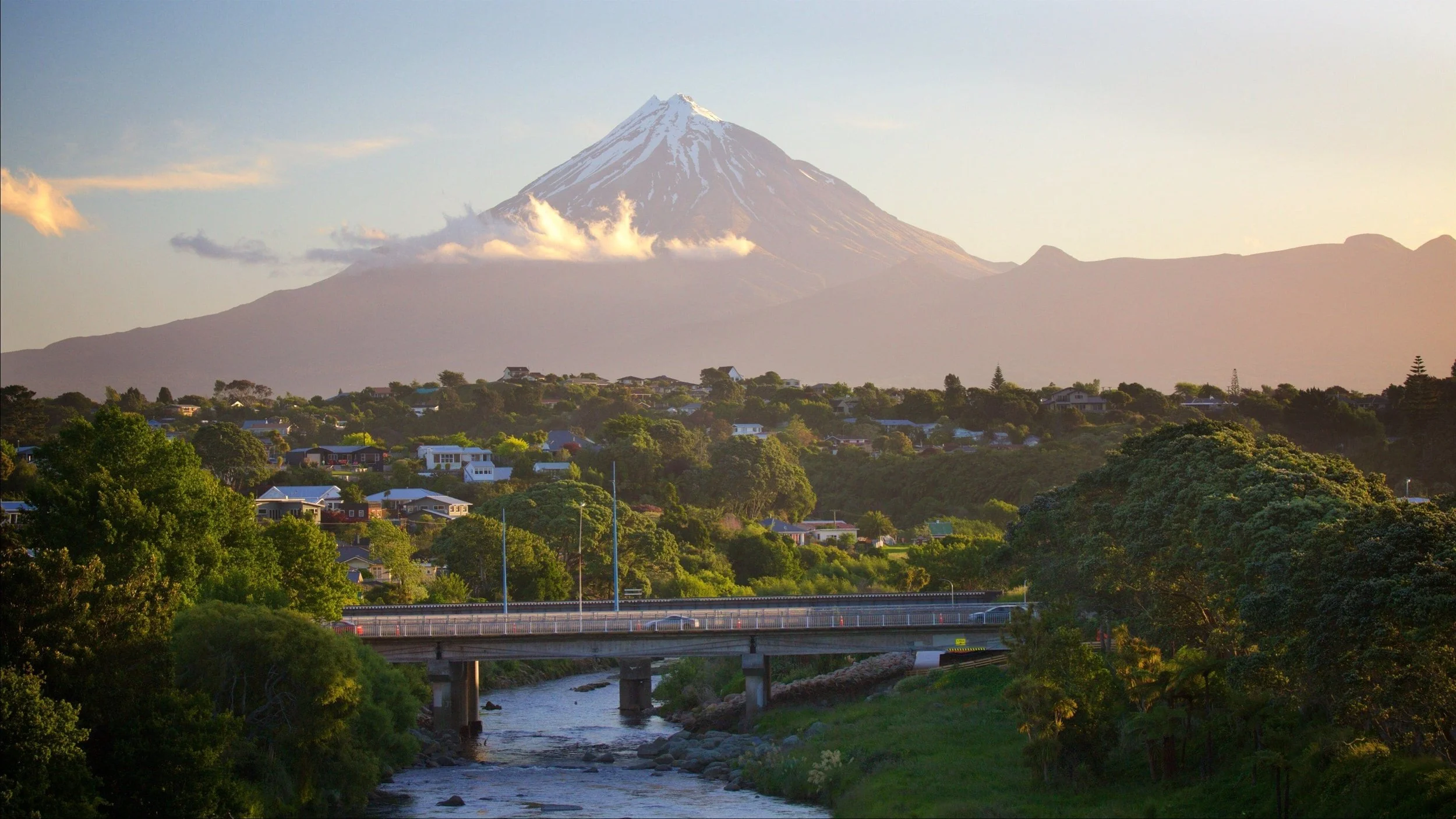 View of Mount Taranaki, also known as Mount Egmont, with snow-capped peak, surrounded by a residential area with houses and greenery, a river in the foreground, and a bridge crossing the river.