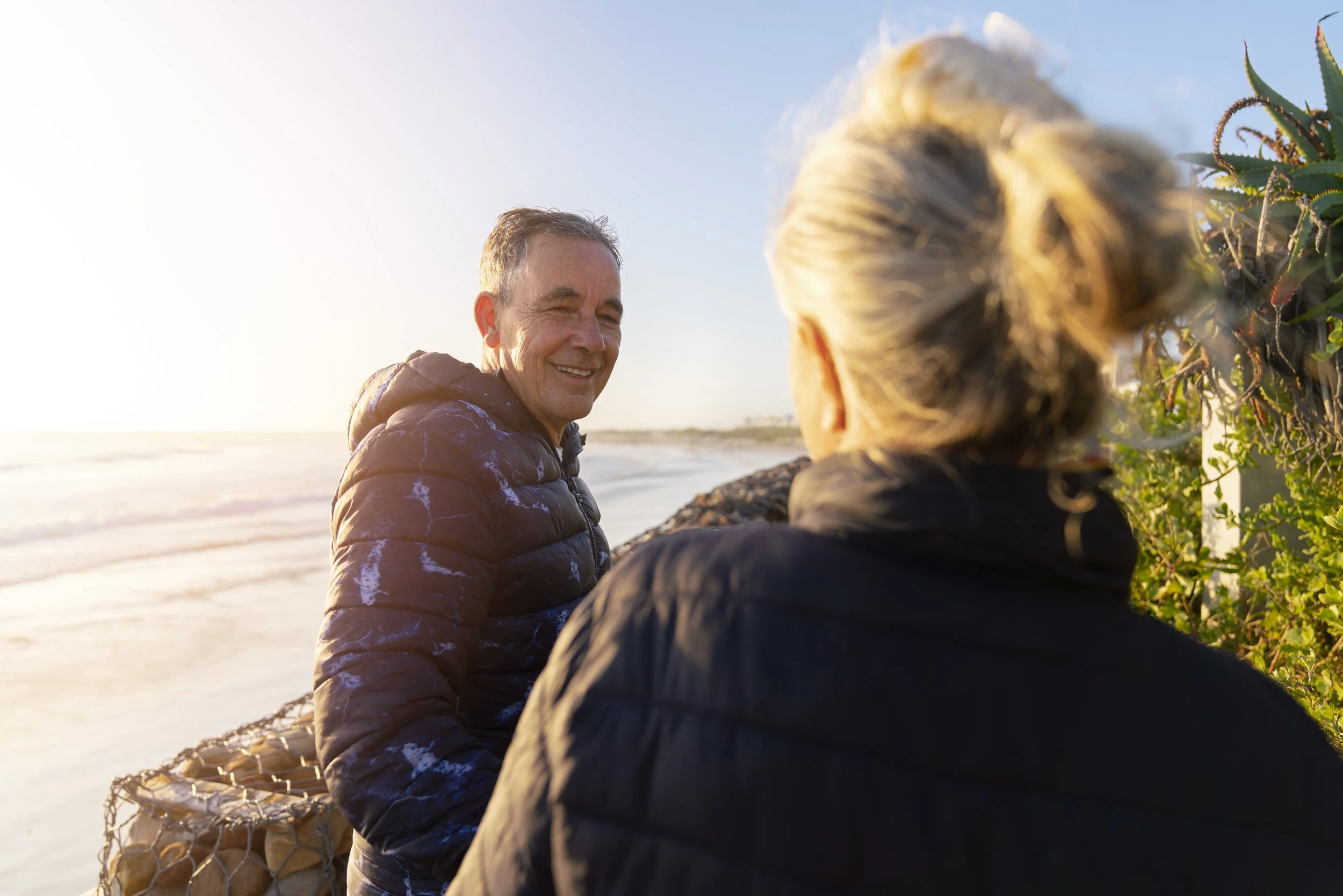 Two people, a man and a woman, standing on the beach near the water, with the man smiling and the woman facing away, during sunset.