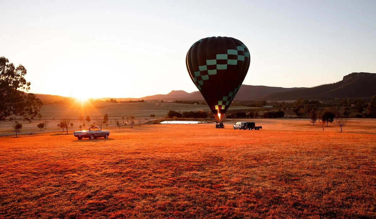 A hot air balloon being prepared for flight during sunrise, with two trucks on a grassy field surrounded by trees and hills.