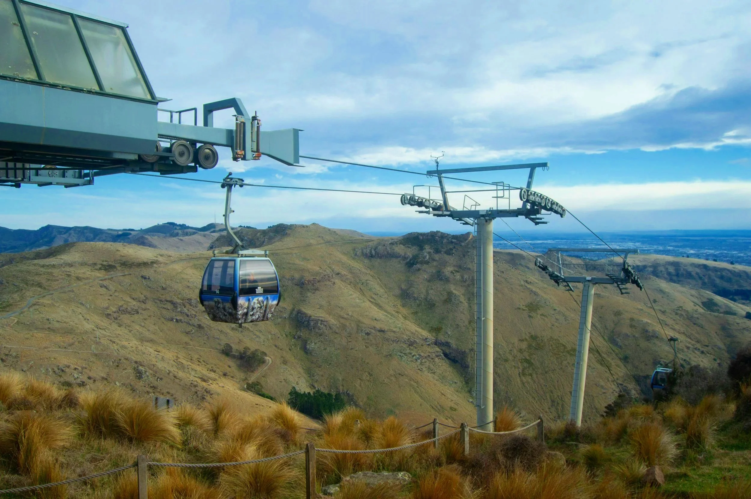 Several gondola lift chairs are traveling over a mountainous landscape with dry grass and sparse vegetation, under a partly cloudy sky.