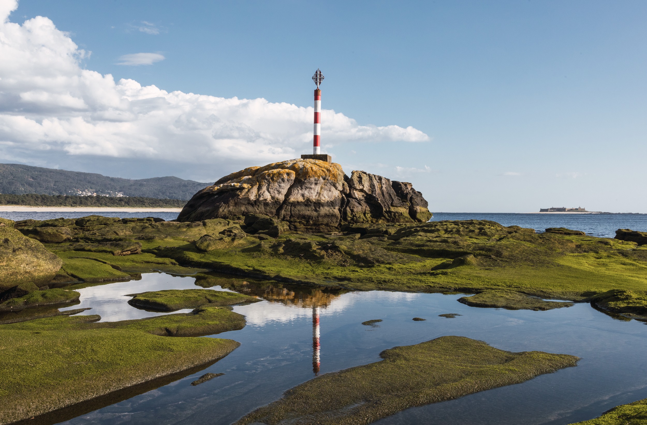 A red and white striped navigation marker pole on a rocky coastal area with green moss, reflecting in a tide pool with water, under a blue sky with white clouds, and distant hills and a ship.