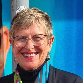 A smiling woman with short gray hair, glasses, and earrings, wearing a black jacket and a multicolored lanyard, standing outdoors in front of a blue background.