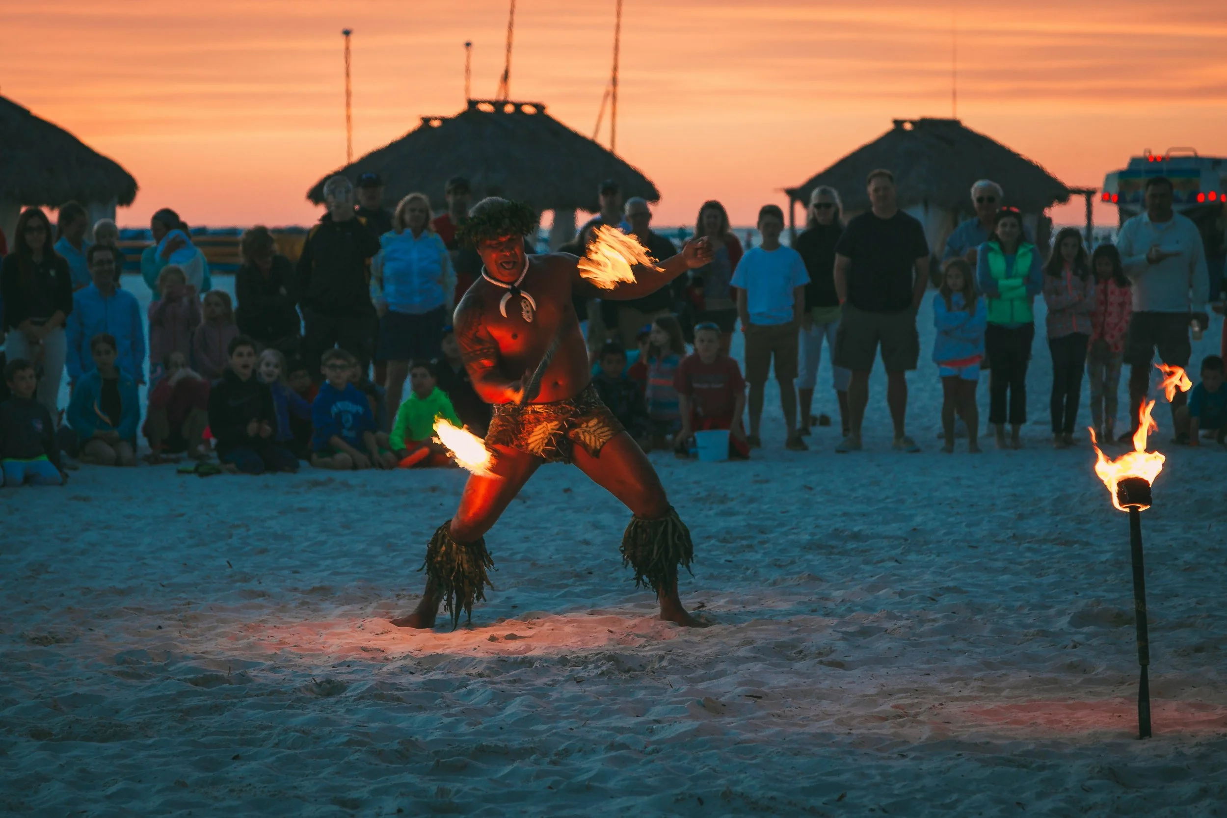A beach scene at sunset with a fire dancer performing in front of a crowd. The performer is holding flaming torches and is surrounded by an audience of adults and children, with thatched-roof huts and a sunset sky in the background.
