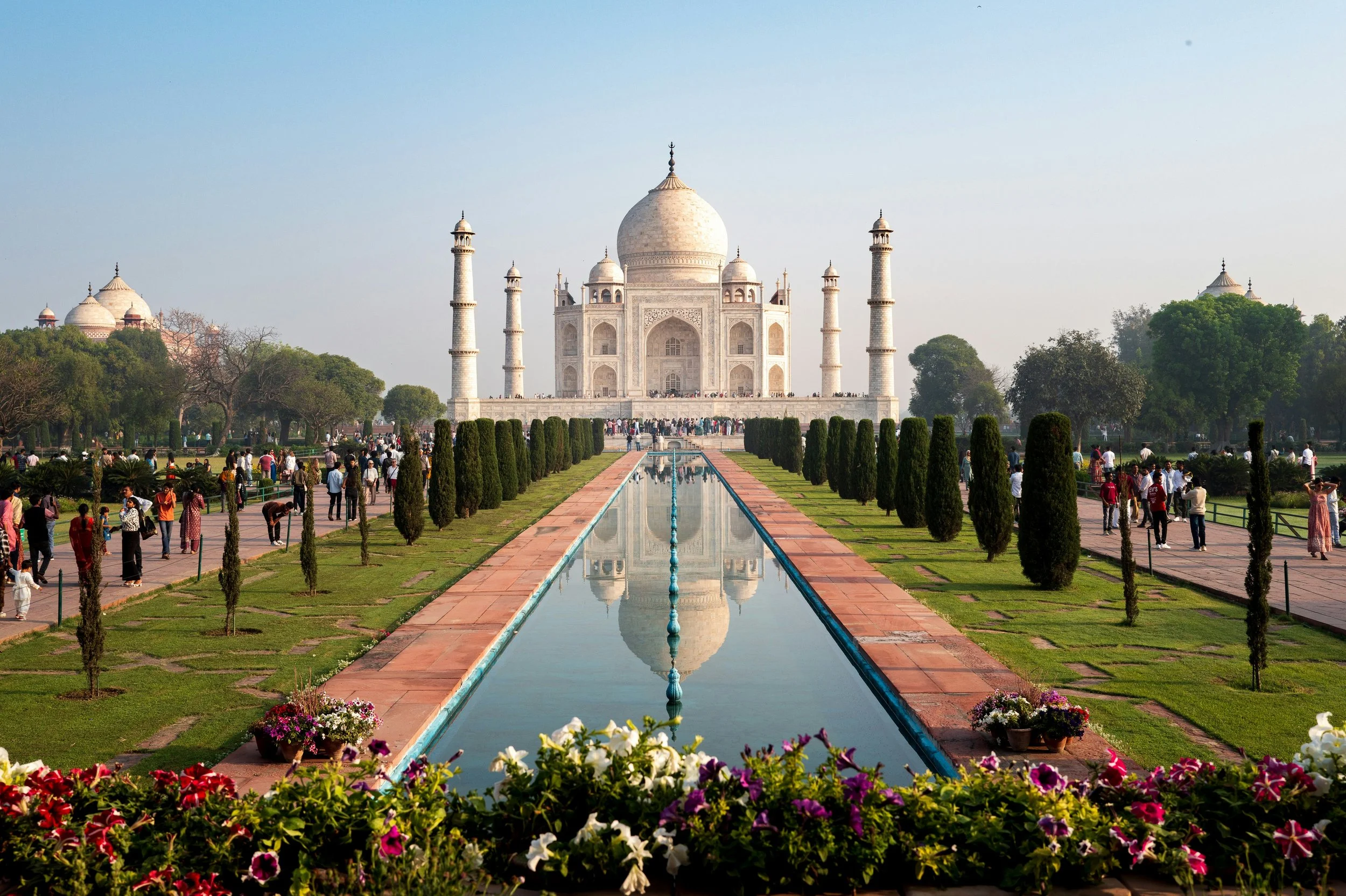 The Taj Mahal, a white marble mausoleum with a large central dome and four minarets, is surrounded by a garden with visitors walking along pathways, trees, and colorful flowers in the foreground, with a reflecting pool leading up to the monument.