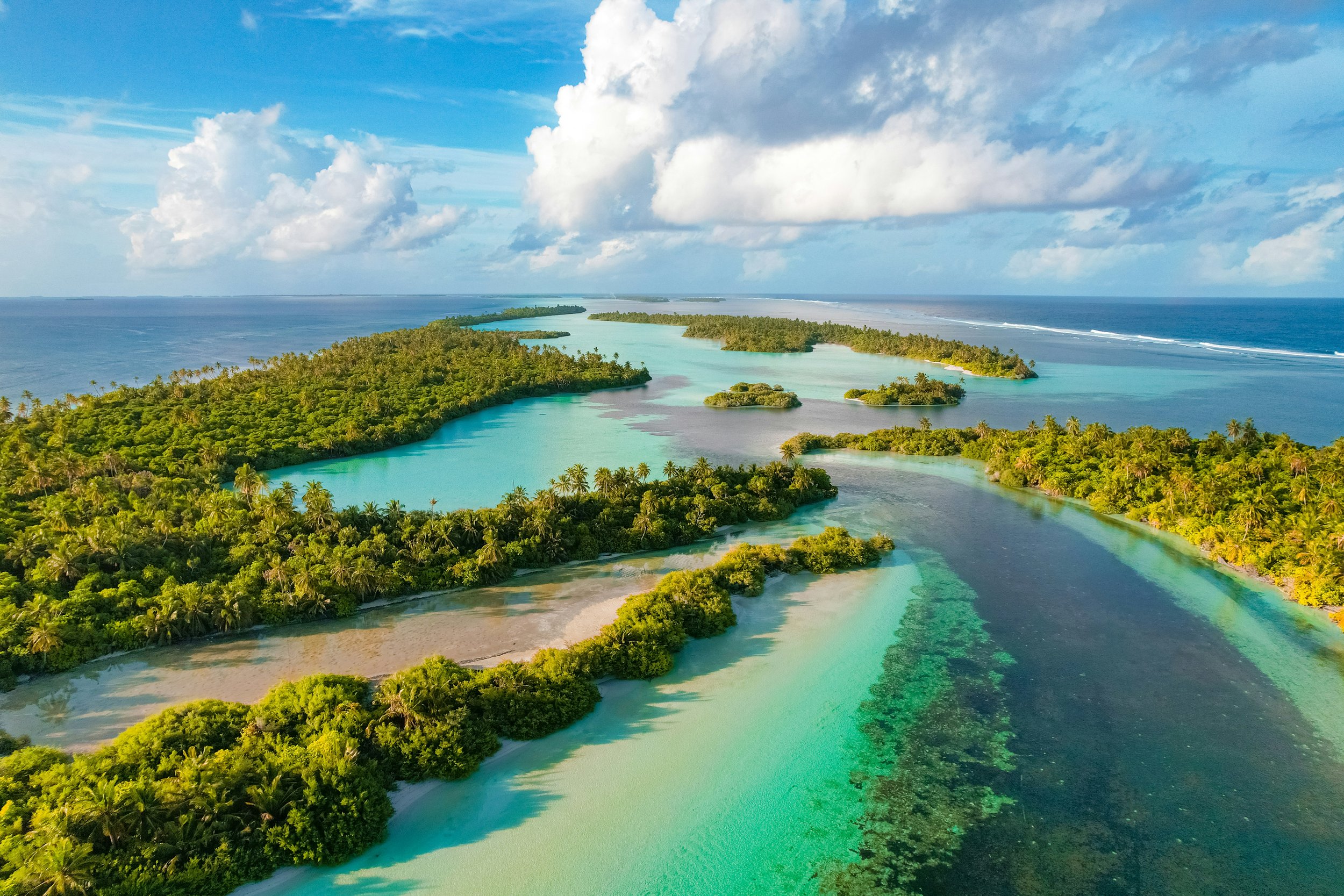 Aerial view of a tropical island with lush green trees, sandy beaches, and turquoise waters, under a partly cloudy sky.