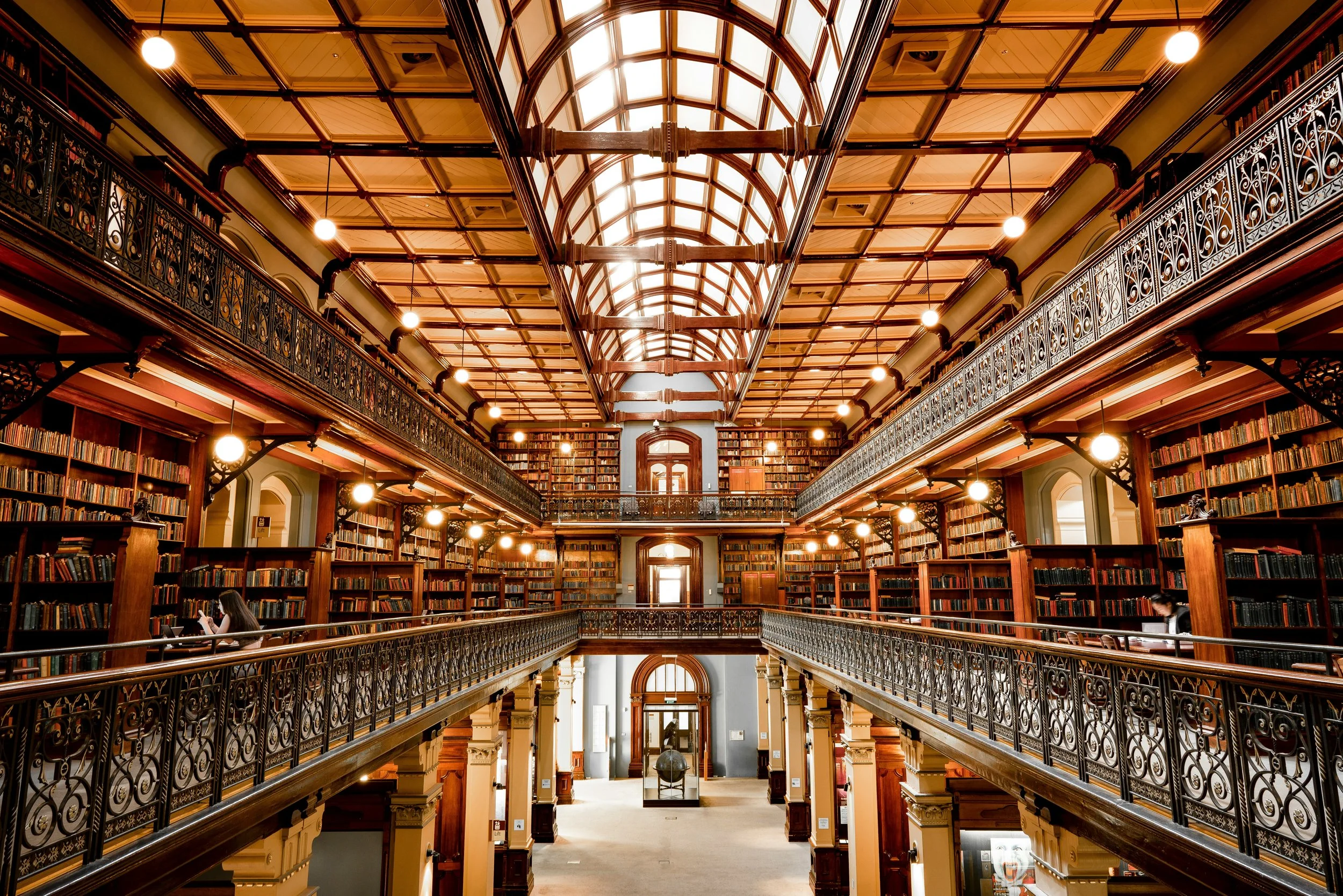 Interior view of a historic library with multiple levels of wooden bookshelves filled with books, ornate iron balconies, and a glass skylight ceiling.