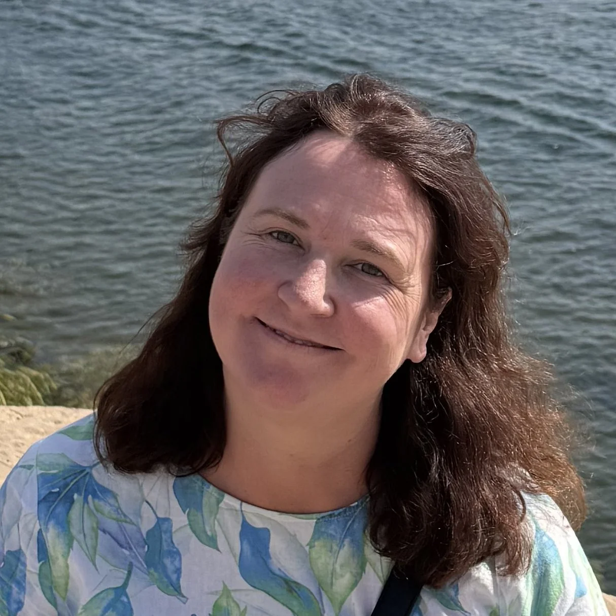 A woman with brown wavy hair smiling near a body of water on a sunny day.