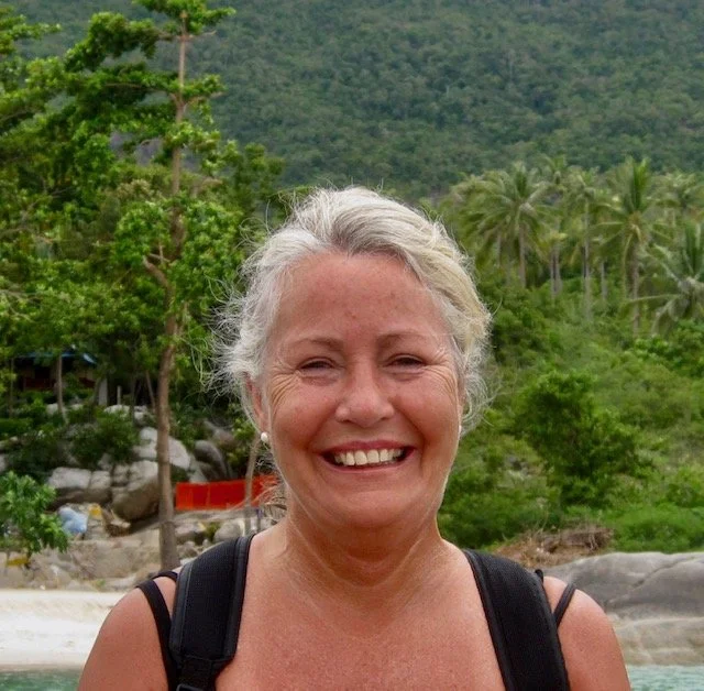Smiling woman with gray hair outdoors in a lush green tropical setting with trees and hills.