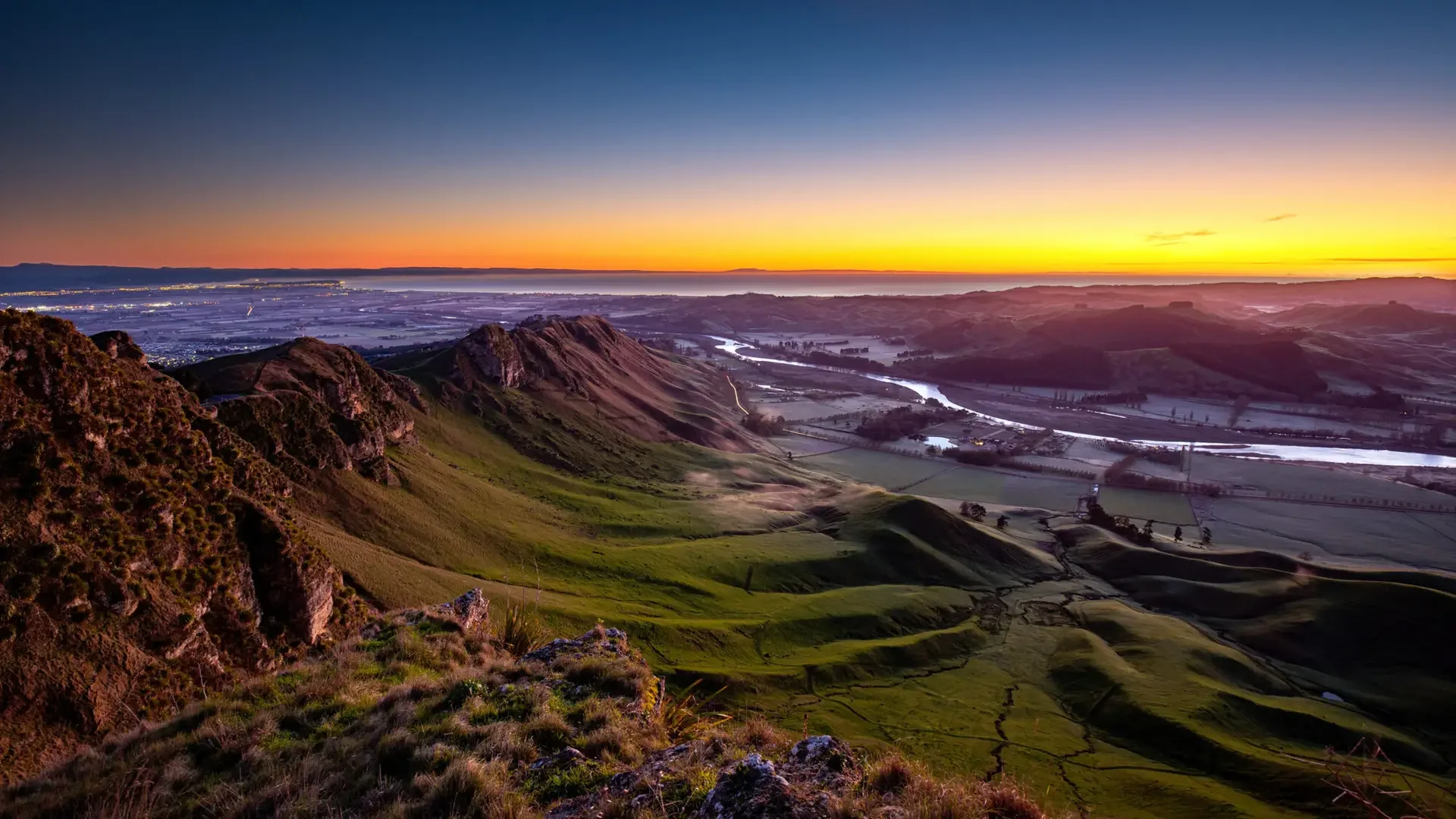 A scenic landscape at sunrise showing rolling green hills, a river winding through the valley, and a vibrant sky with orange and blue hues.