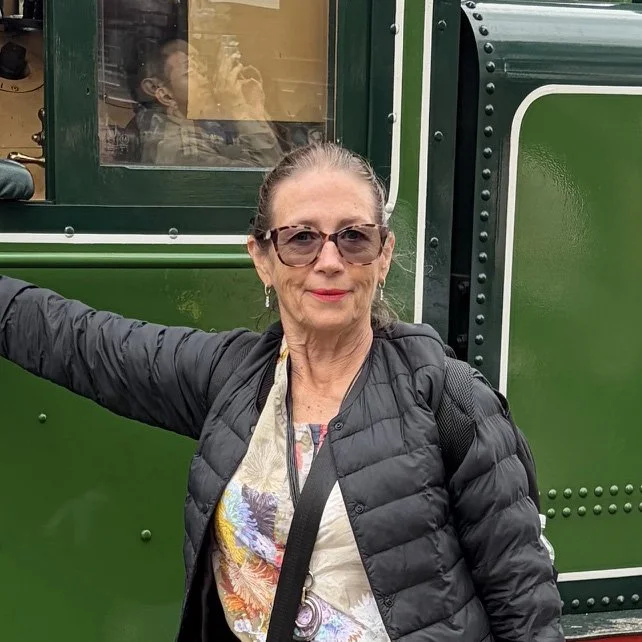 A woman with glasses and a black puffer jacket standing in front of a green train, with a passenger visible inside the train behind a window.