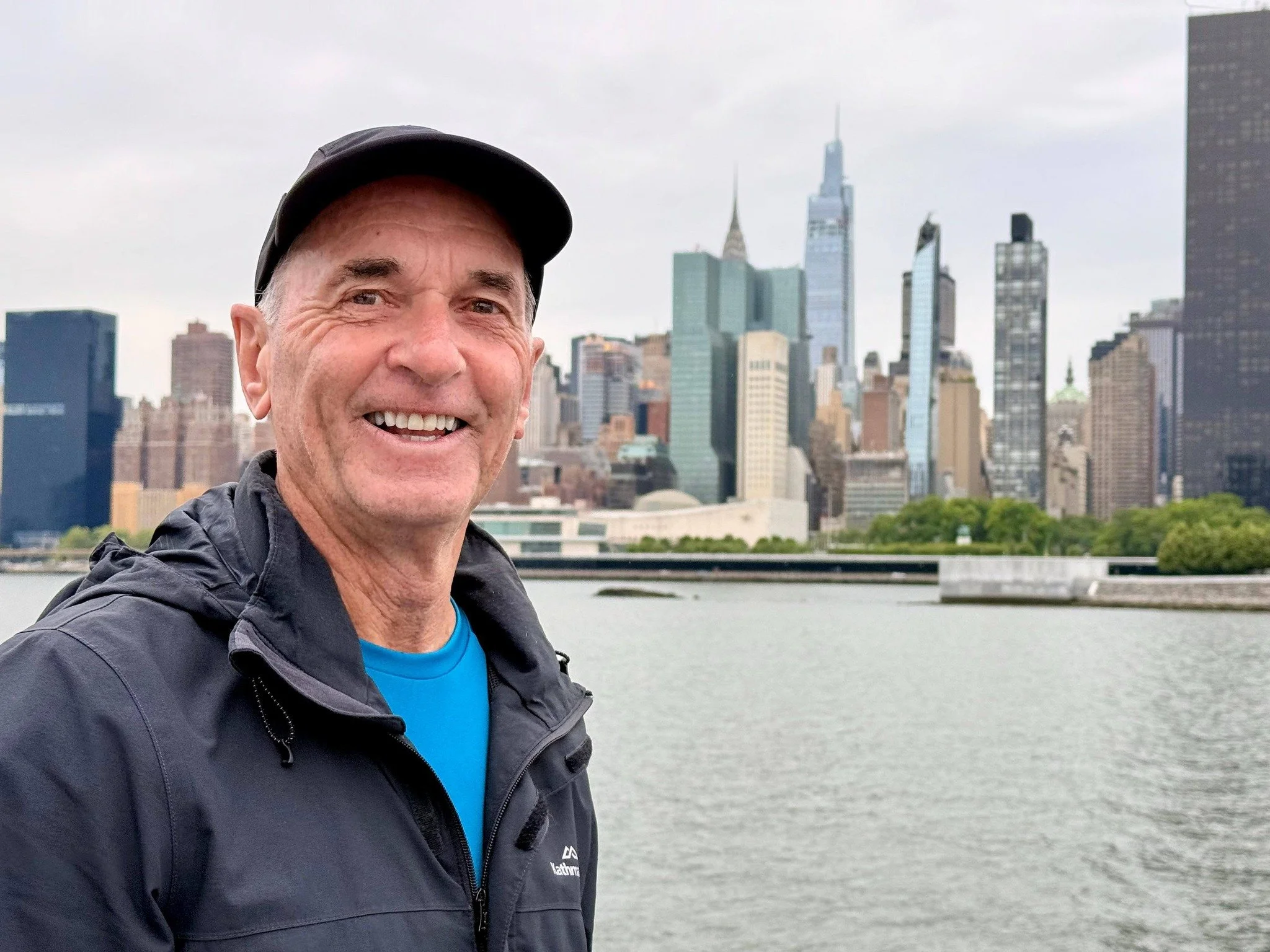 Smiling man in outdoors with city skyline in background, including tall skyscrapers and a river in the foreground.