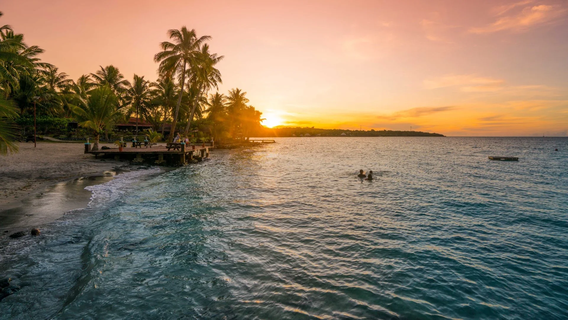 Sunset over a beach with palm trees, a pier, and people swimming.