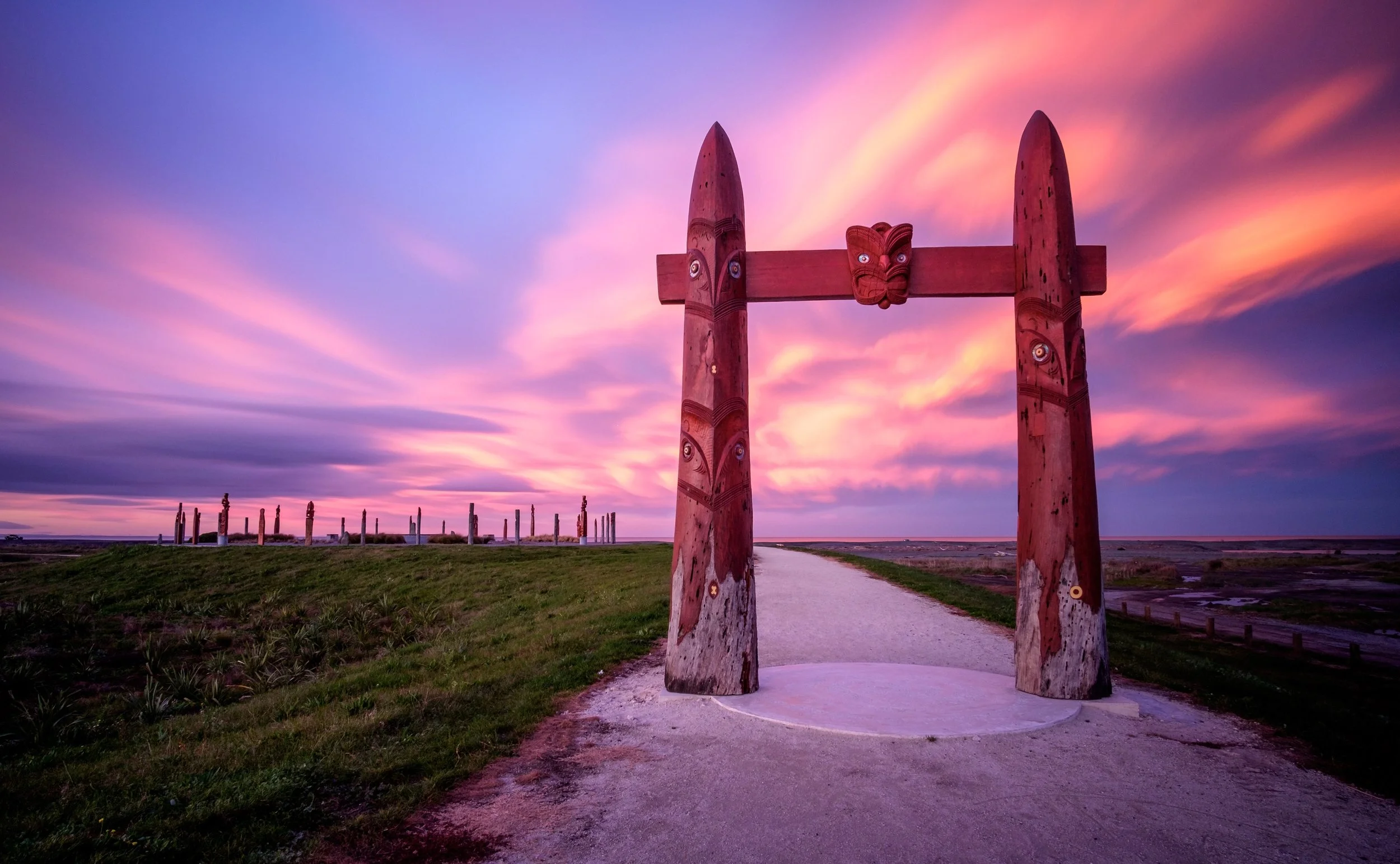 A scenic landscape with a wooden archway featuring carved figures, set against a colorful sunset sky with pink and purple clouds, and a pathway leading toward more carved wooden posts in the distance.