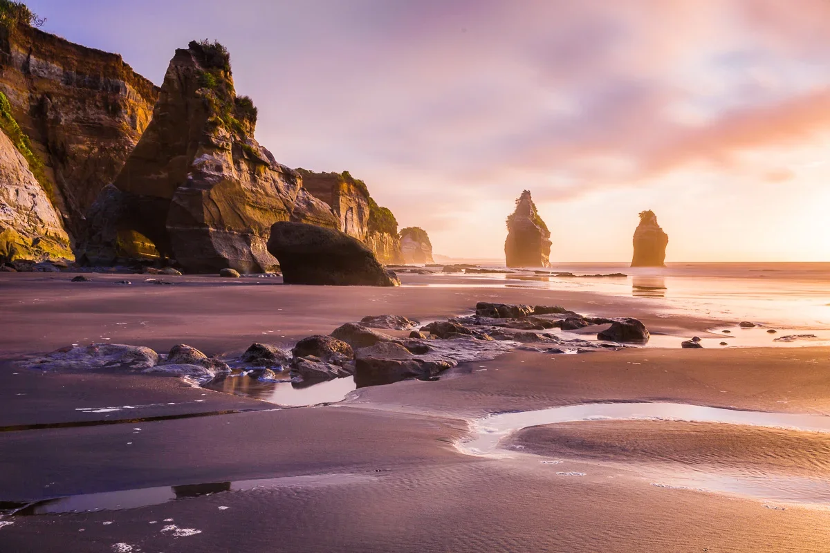 Sunset over a beach with large rock formations and cliffs in the background.
