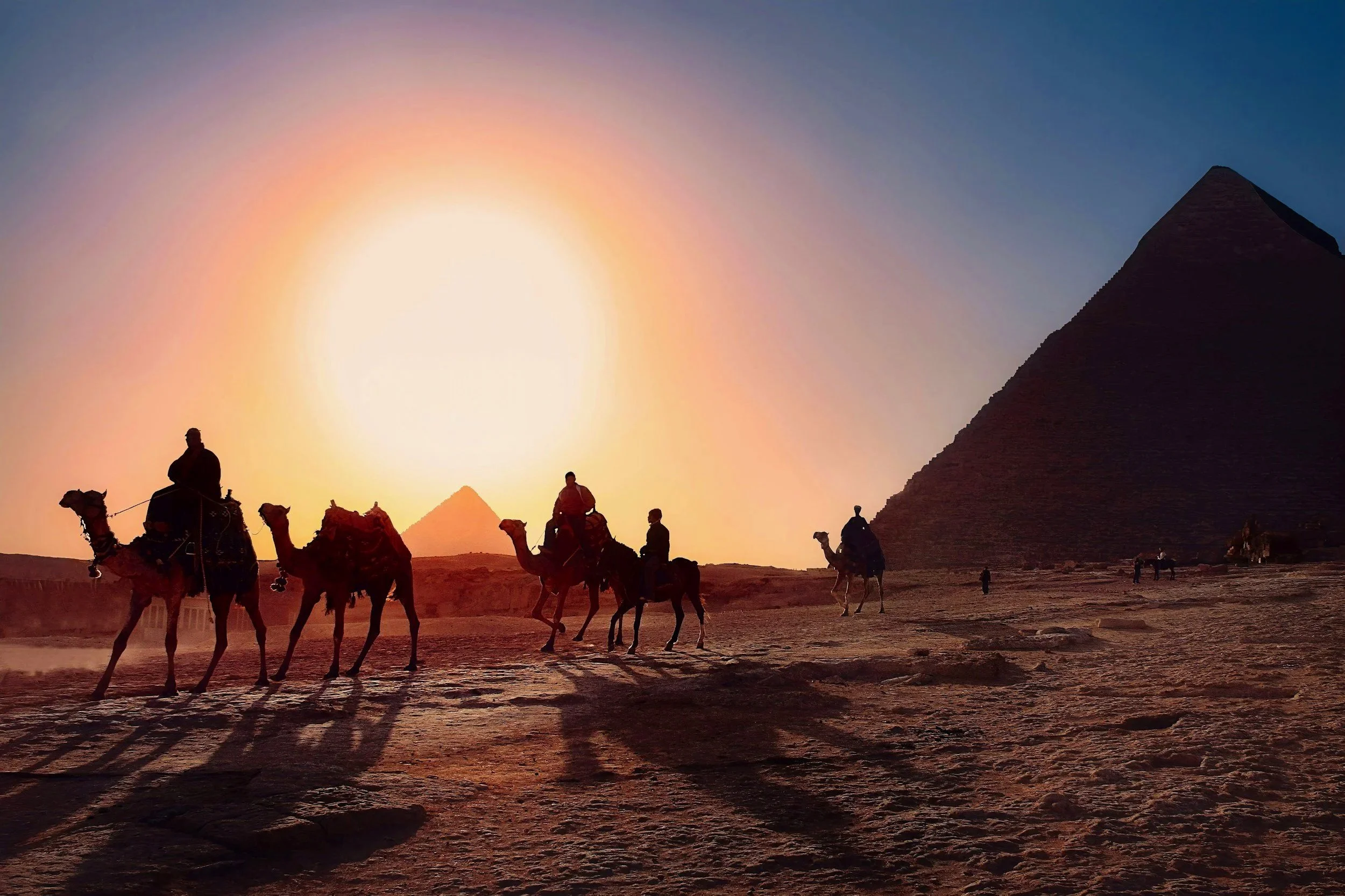 Silhouettes of people riding camels in front of the pyramids during a sunset.
