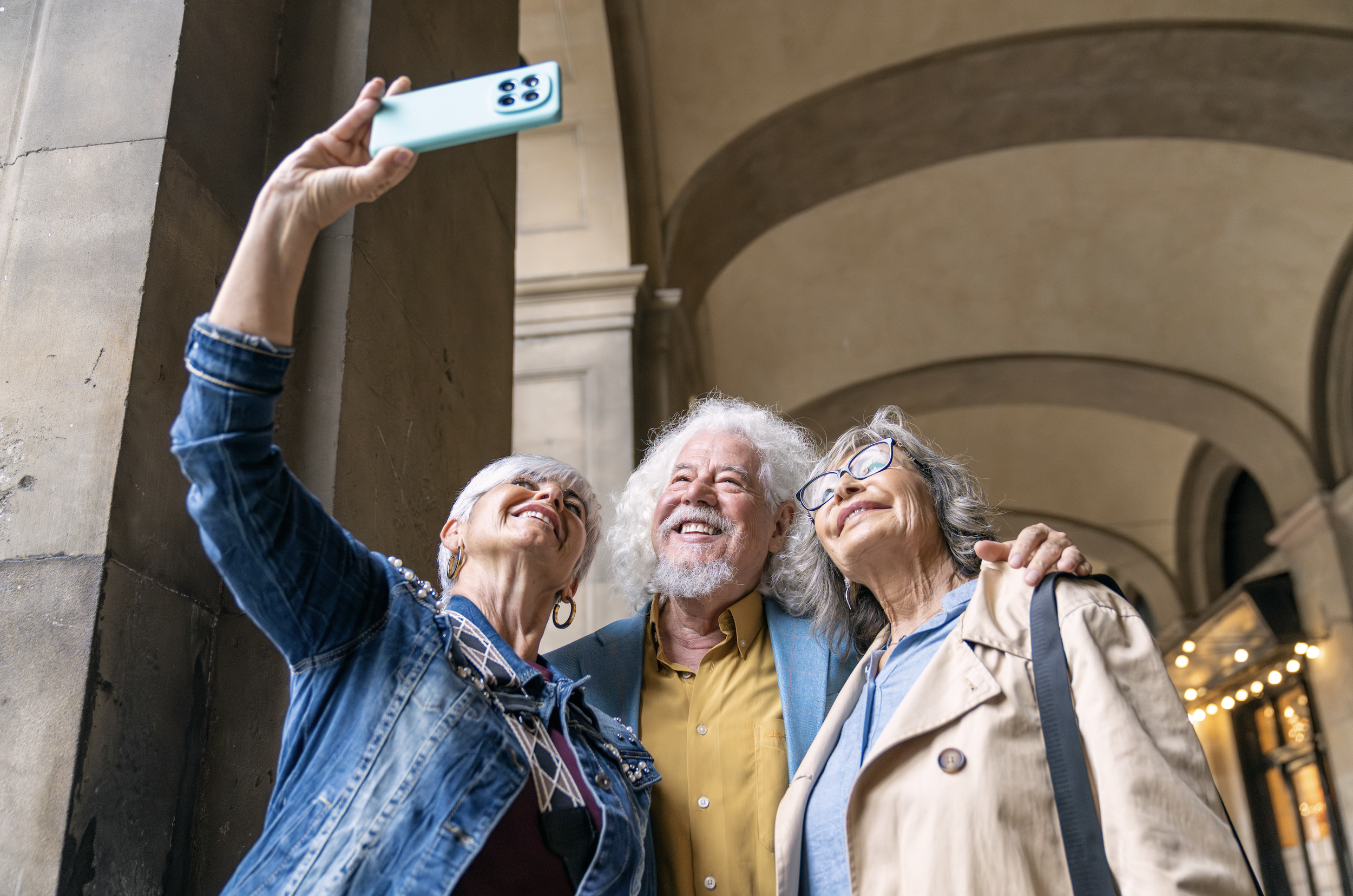 Four elderly people, two women and two men, taking a selfie together in an outdoor covered area, smiling and posing closely.