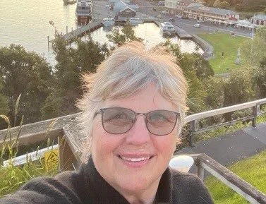 A woman with short blonde hair and glasses smiling outdoors, with a river, trees, and buildings in the background.