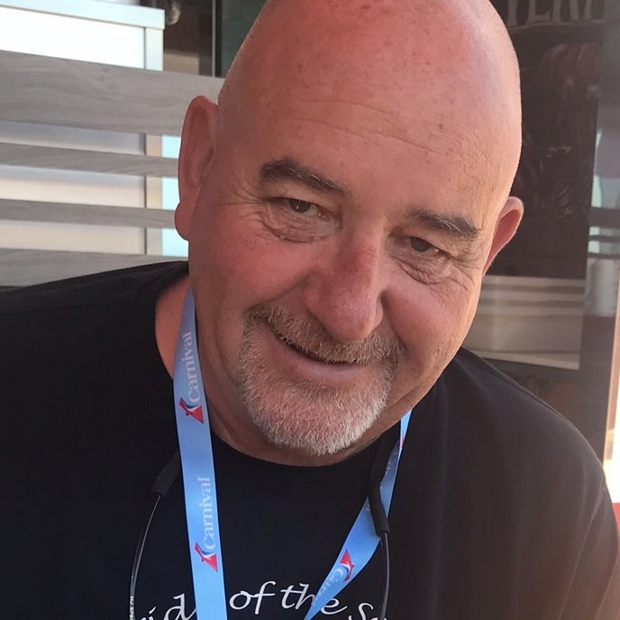 A smiling, bald man with a goatee and a black shirt, wearing a blue lanyard, sitting outdoors near a building.