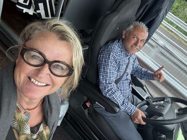 A woman and a man smiling for a selfie; the man is sitting in the driver's seat of a truck, pointing out the window, while the woman is standing outside, taking the photo.