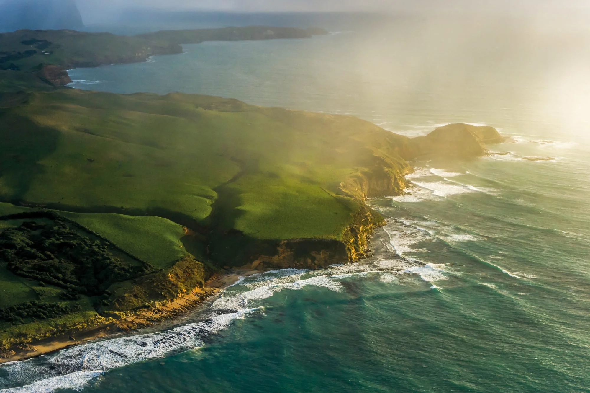 Aerial view of lush green coastal cliffs with waves crashing on the shoreline during sunset.