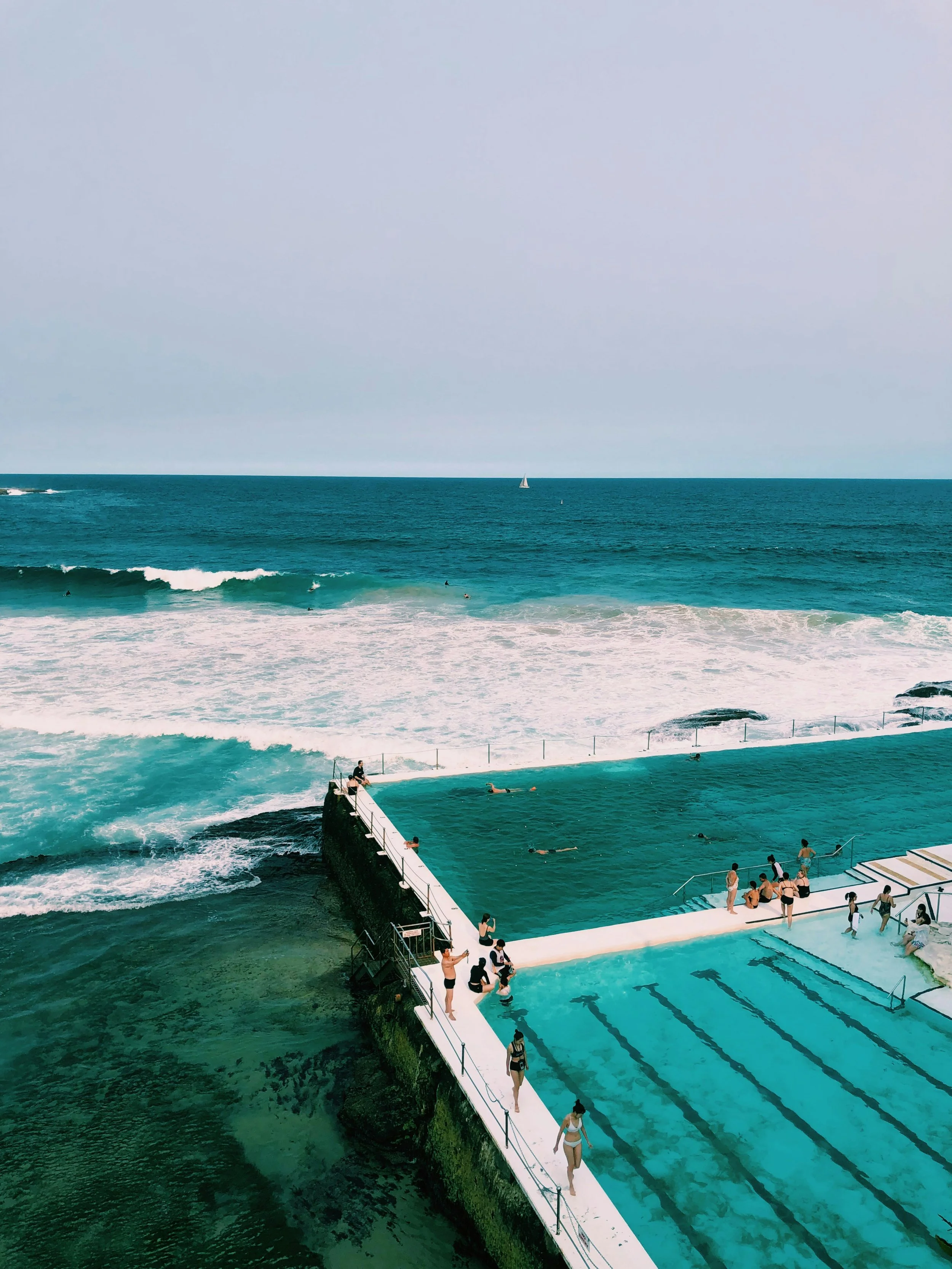 People swimming and relaxing in an ocean pool with an ocean view, near the coast, with a sailboat and a yacht on the horizon.