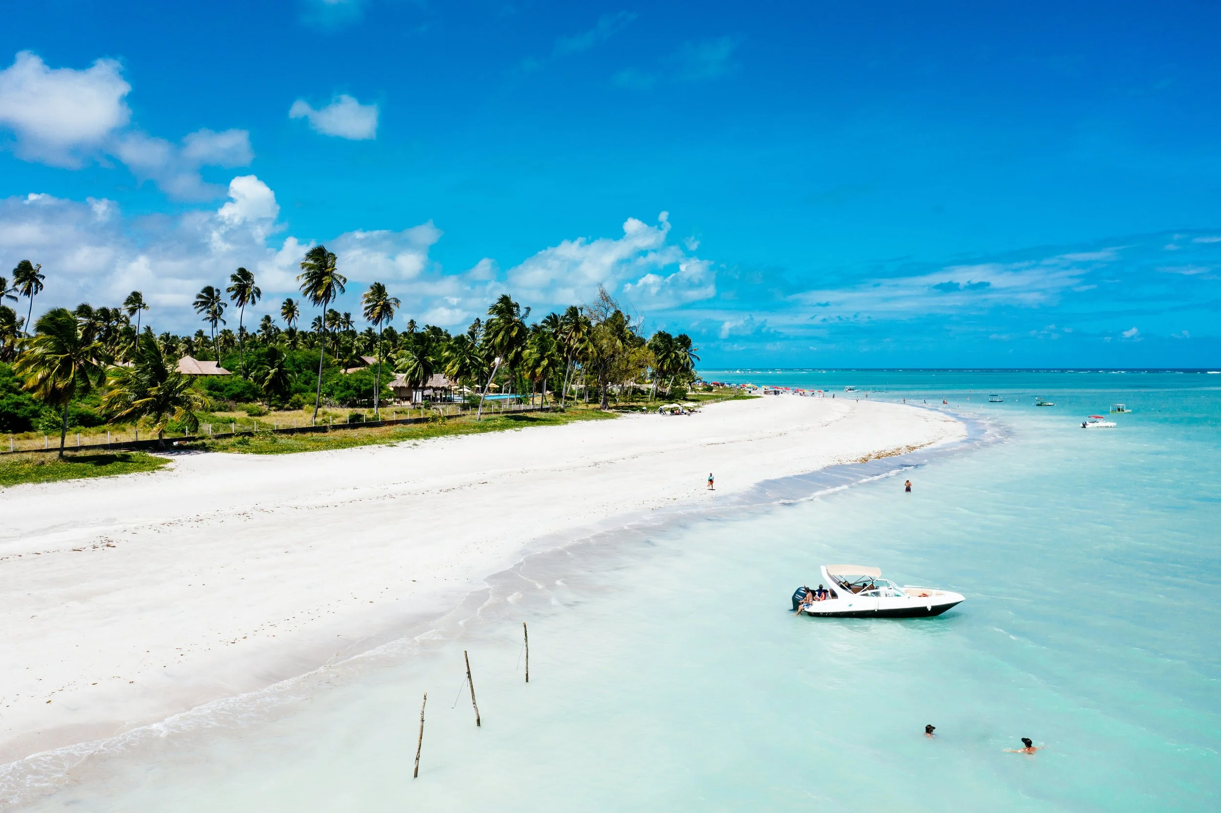 Tropical beach with white sand, palm trees, blue sky with a few clouds, and clear water with boats and people enjoying the beach.