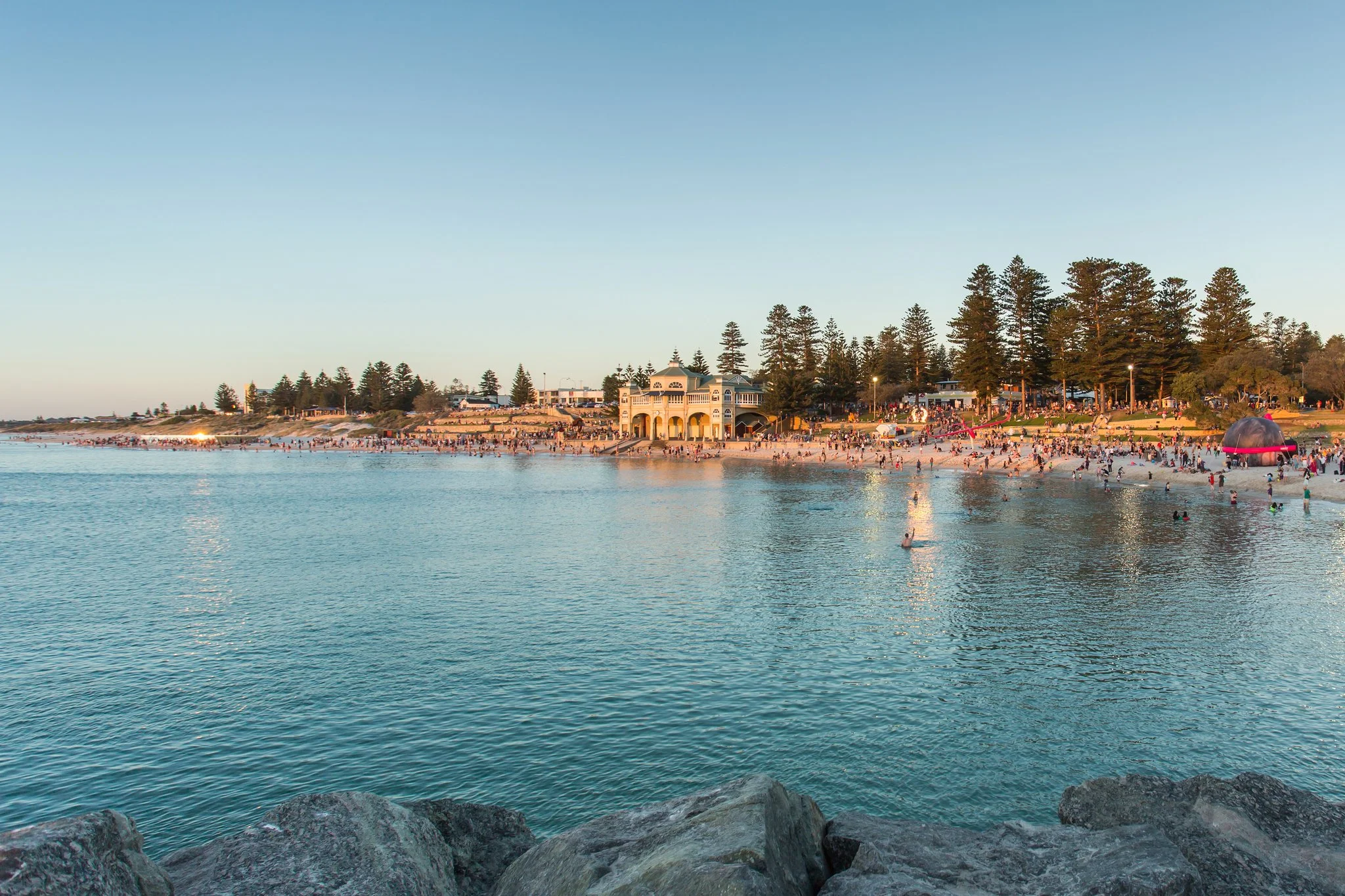 A crowded beach with many people swimming and sunbathing near a large, ornate building across the shoreline, with a clear blue sky overhead.