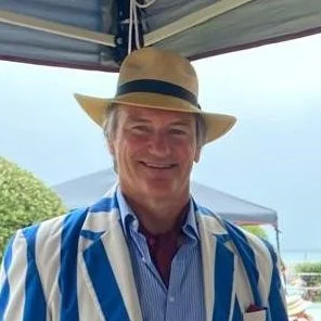A smiling man wearing a straw hat and a blue and white striped blazer, outdoors near a lake or ocean.