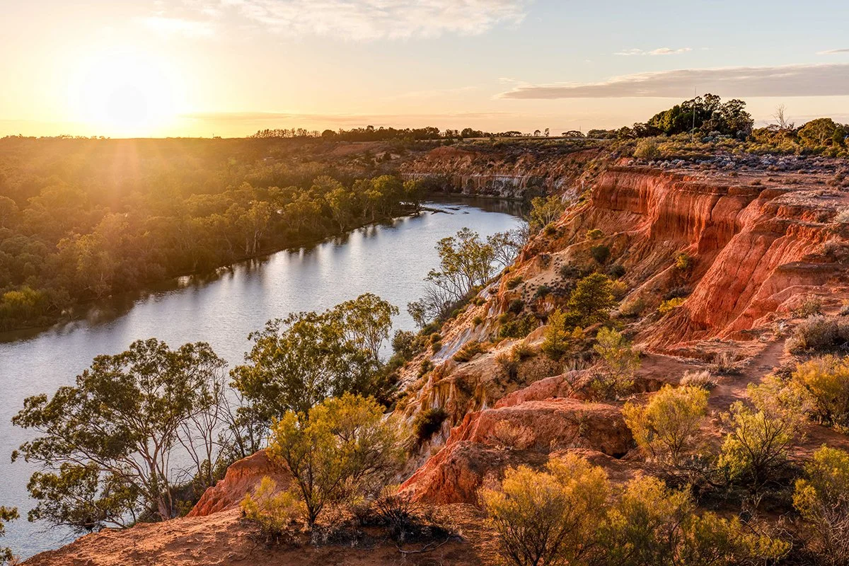 Sunset over a river flowing through a canyon with red and orange cliffs and sparse green shrubs and trees.
