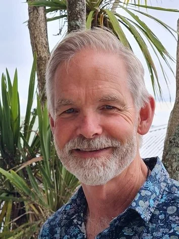 A smiling man with gray hair and beard wearing a blue patterned shirt outdoors near palm trees.