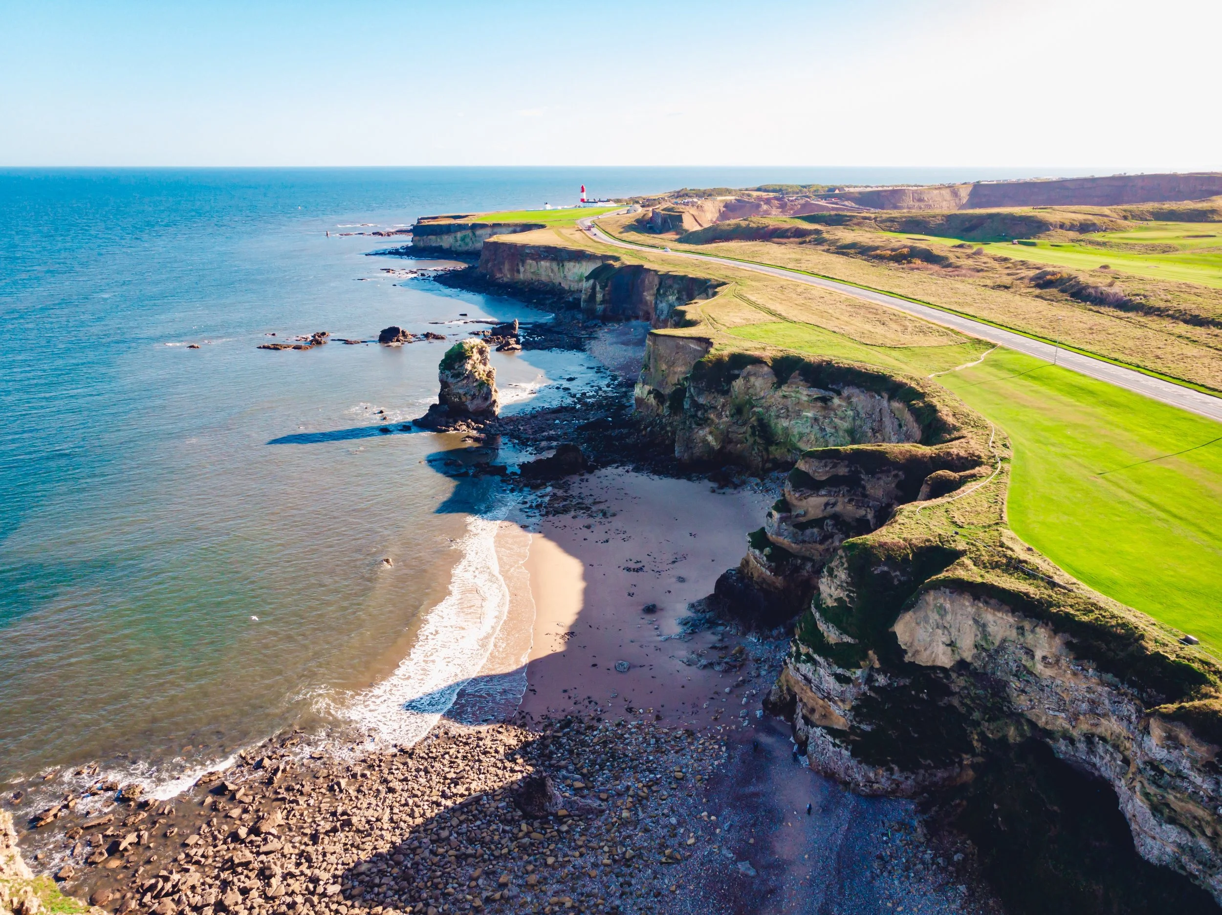 Coastal cliffs with a lighthouse in the background, overlooking the ocean and a sandy beach below.