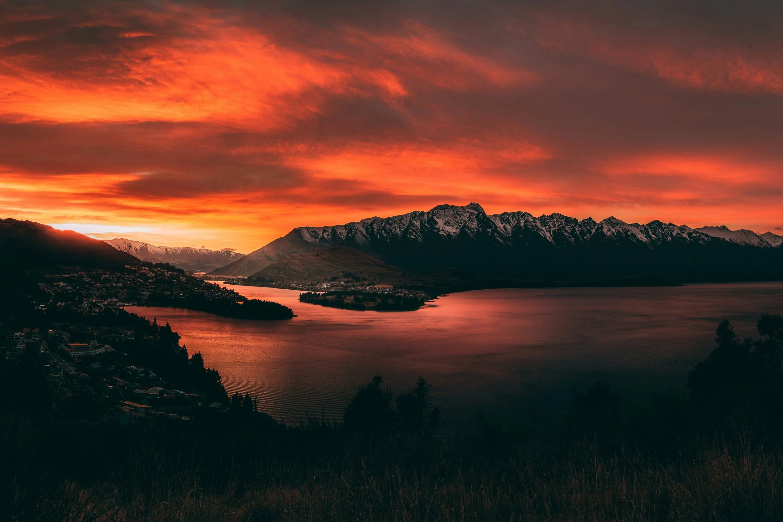 A scenic view of a sunset over a mountain range with snow-capped peaks, a lake in the foreground, and a town along the shoreline, with a colorful sky filled with orange and pink clouds.