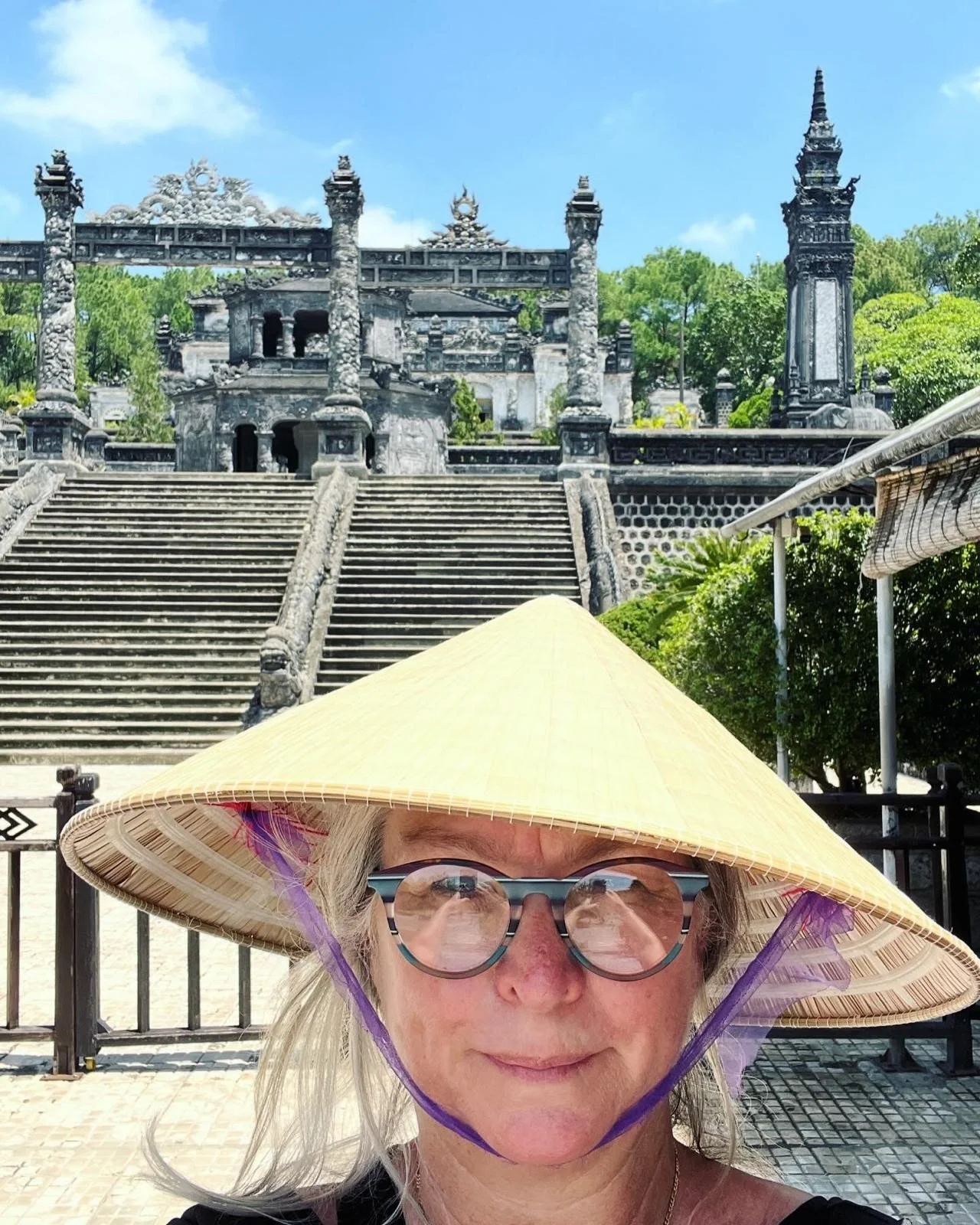 A woman with glasses and a wide-brimmed straw hat taking a selfie in front of an ancient Asian temple with stone steps and intricate architectural details, surrounded by green trees.