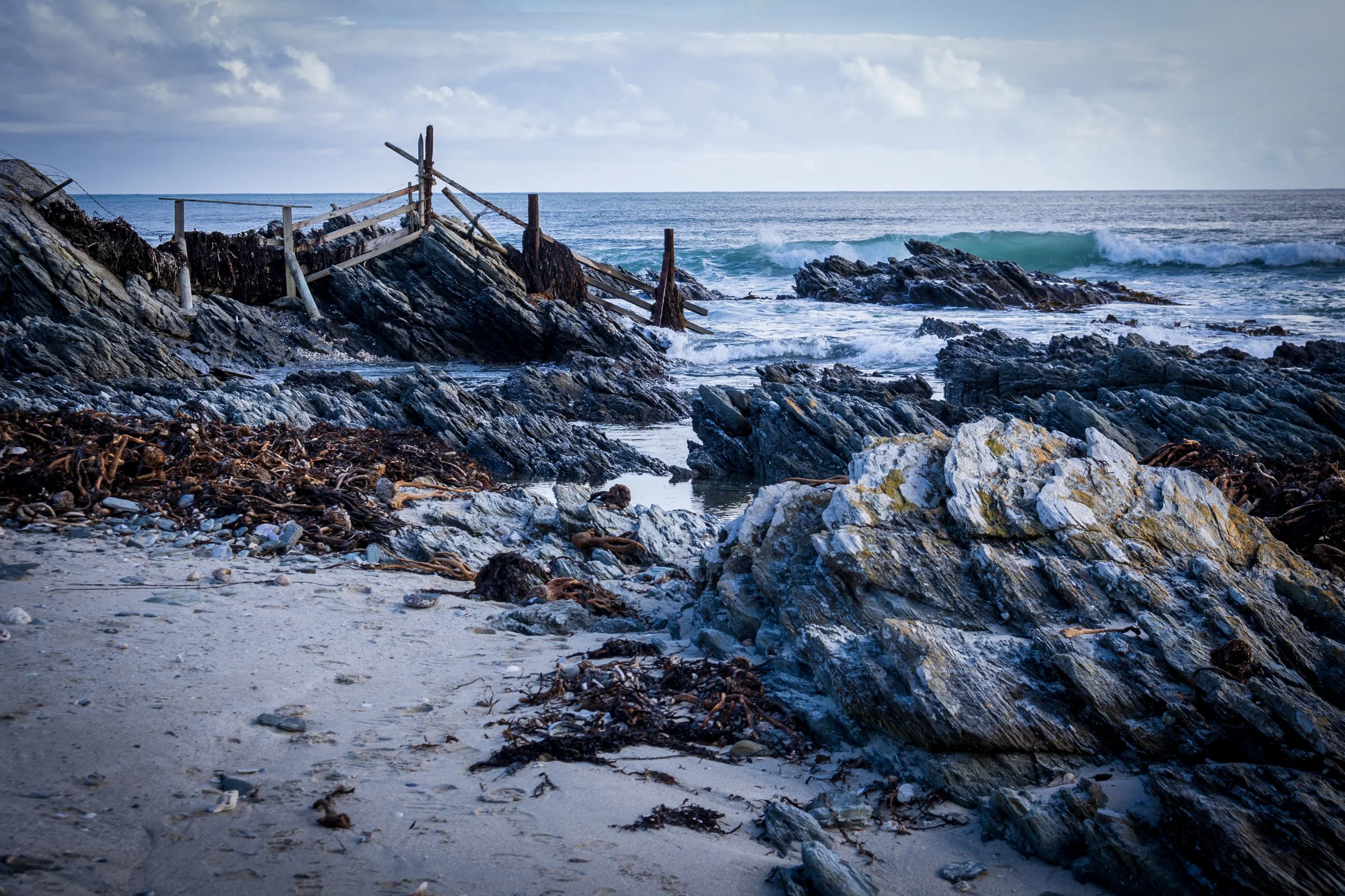 A rugged beach with rocks, seaweed, and driftwood, with waves crashing against the rocky shore under a cloudy sky.