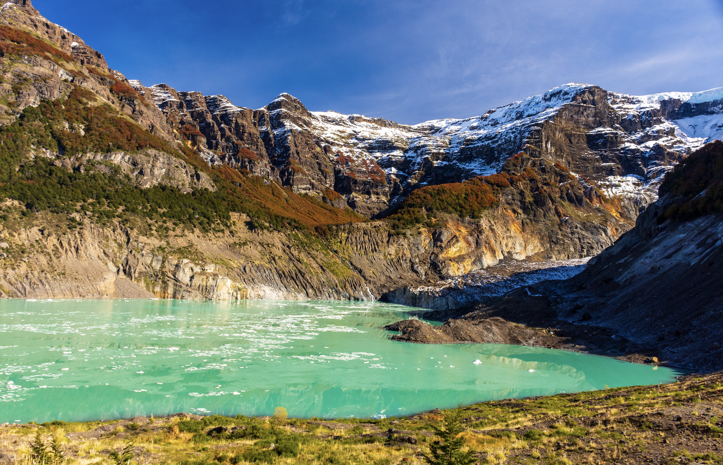 Snow-capped mountains with a steep rocky incline, green and reddish vegetation, and a body of vibrant turquoise water in the foreground beneath a clear blue sky.