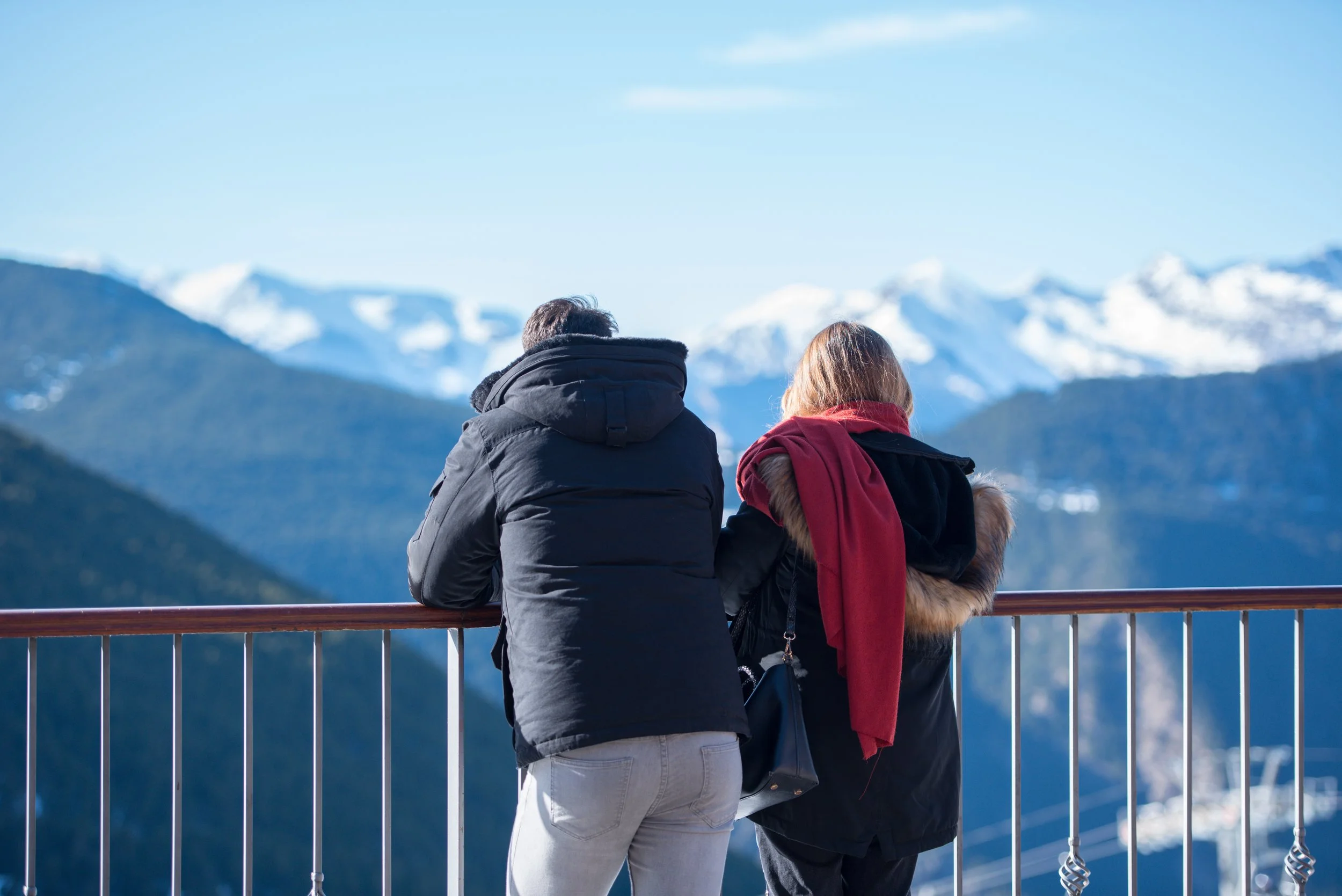 Two people, a man and a woman, stand at a railing looking at snow-capped mountains in the distance, dressed in winter clothing.