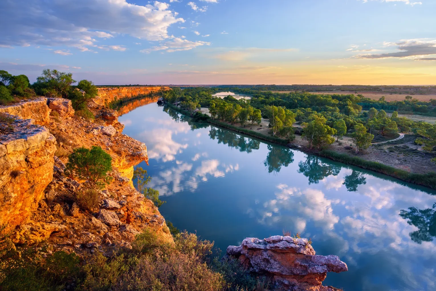 A scenic view of a river reflecting the sky and clouds, flanked by rocky cliffs and lush green trees, during sunset.
