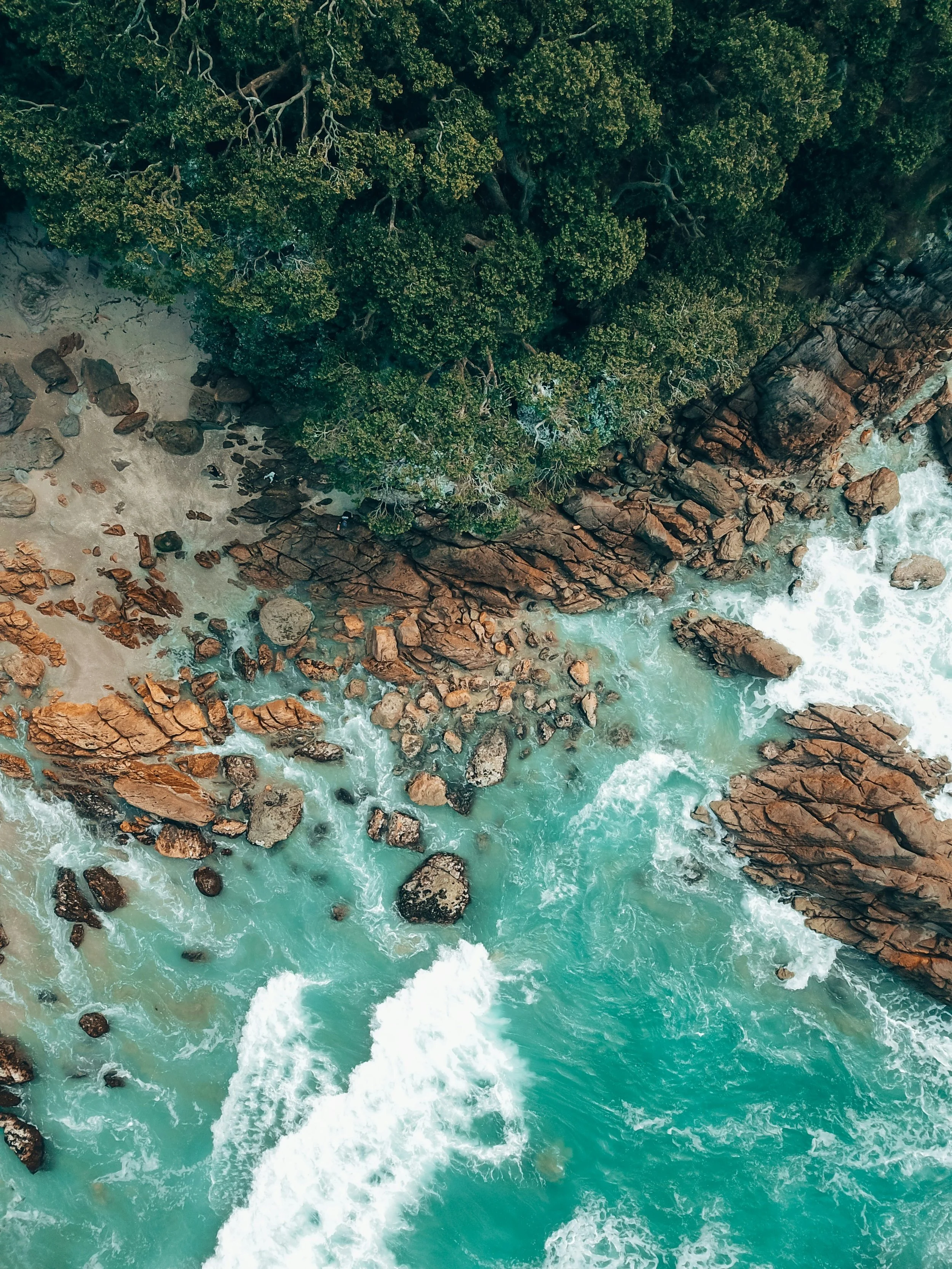 An aerial view of a coastline with large rocks, turquoise water, and a dense green forest.