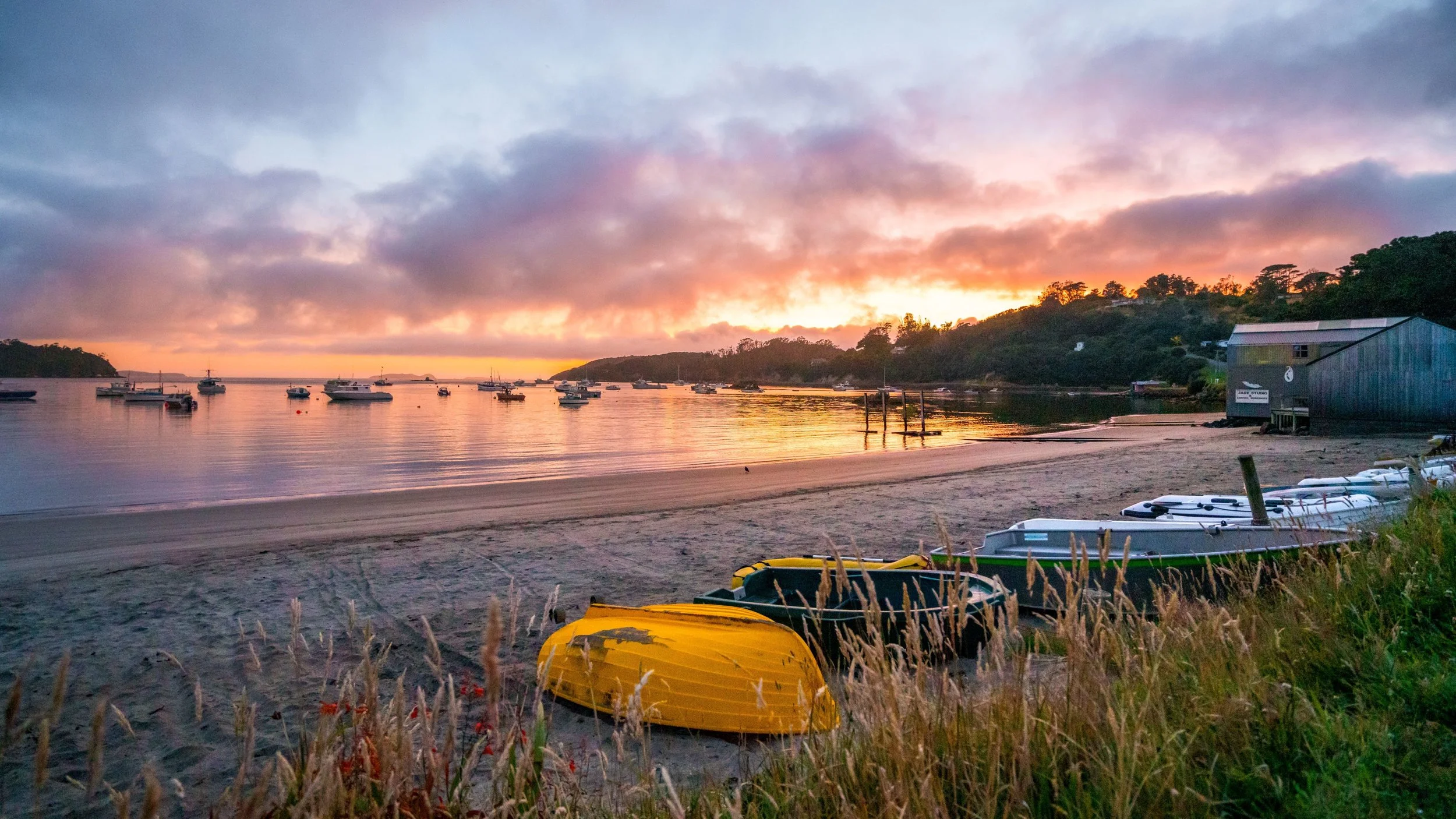 A peaceful harbor scene at sunset with boats anchored in the water, a sandy beach with kayaks, and a small building on the right. The sky is colorful with clouds and the water reflects the sunset colors.