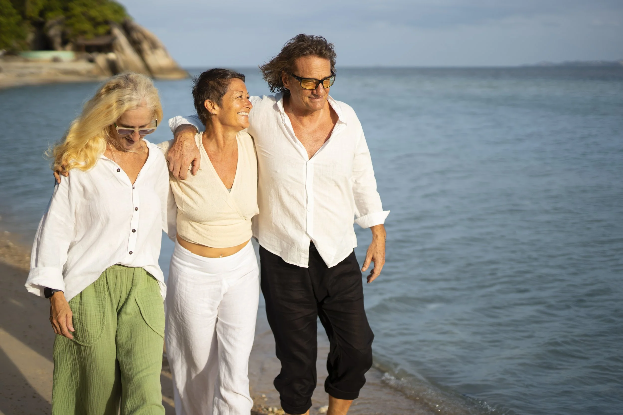 Three older adults walking along the beach near the sea, smiling and enjoying a pleasant day.