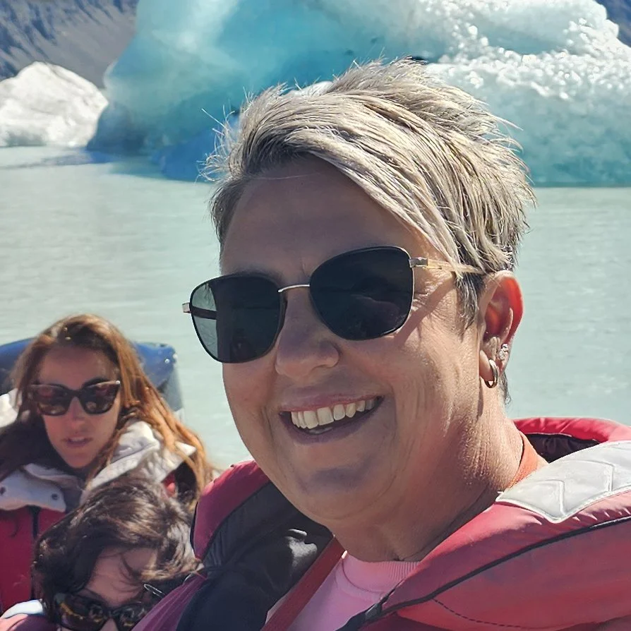 Smiling woman with short blond hair, wearing black sunglasses and a pink jacket, on a boat with icy glacier in the background, accompanied by two women and a dog.